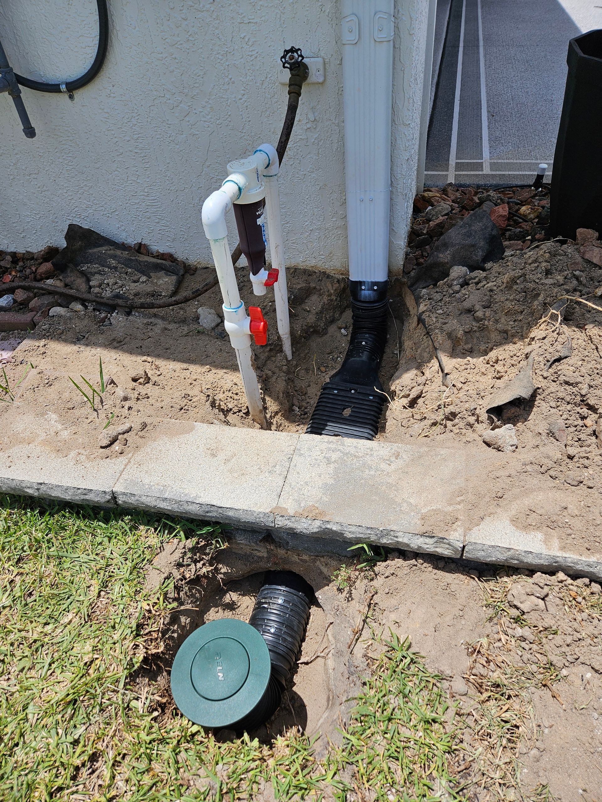 A white PVC pipe system with a red valve and a black drainpipe, next to a gutter and a lawn with a drain.