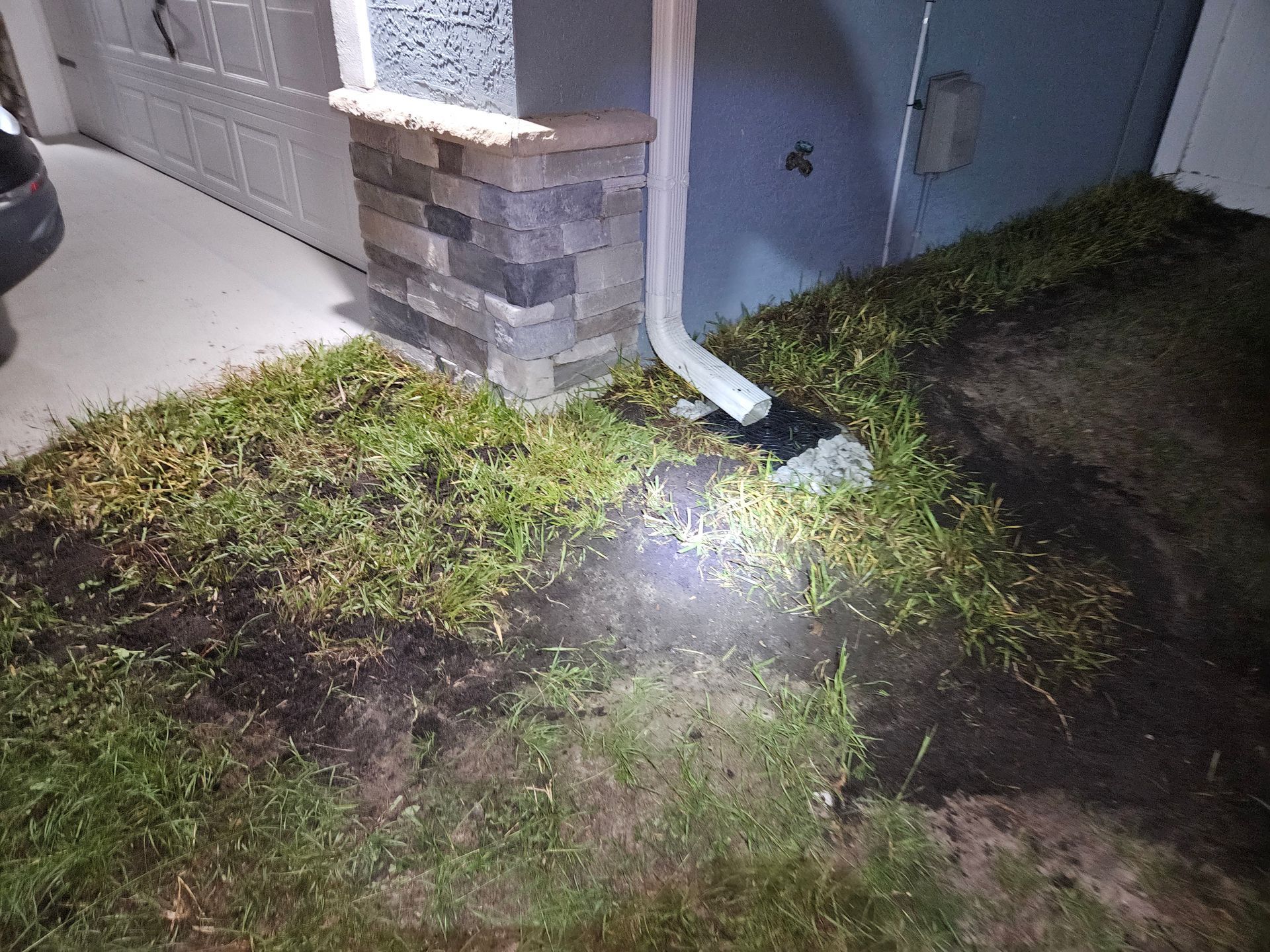 Downspout drains onto lawn near garage. Wet grass and dark soil, possibly after rain.