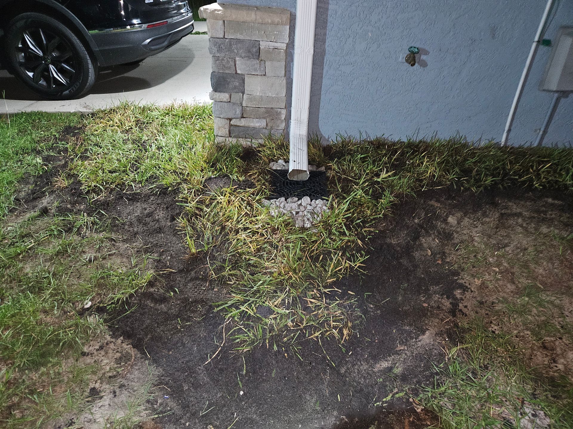 Downspout drains into a dark, muddy area with sparse, yellowing grass; a car is in the background.