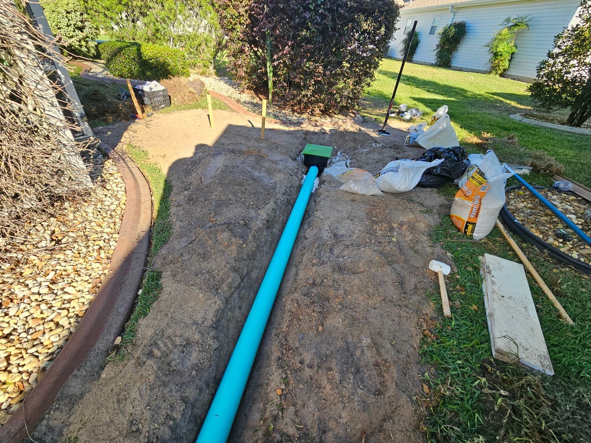 Green pipe and drain in a dirt yard, tools and bags nearby; residential garden setting.