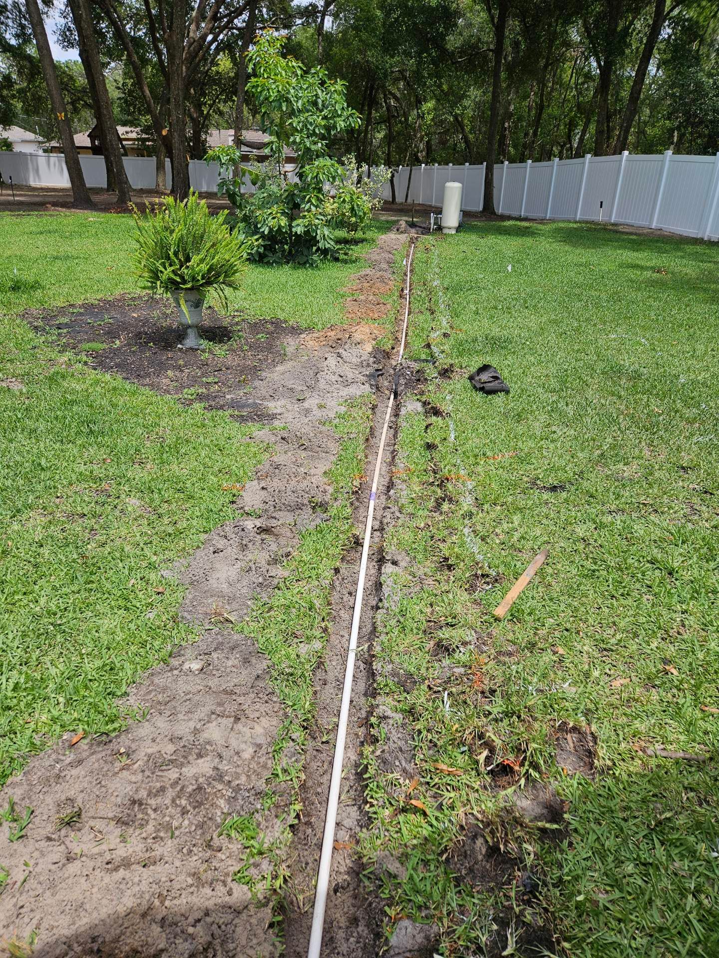 A trench dug in a grassy yard with a white pipe inside, near a white fence and trees.