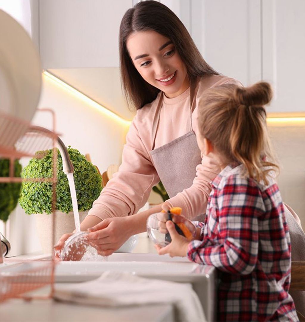 Woman and child washing hands at a kitchen sink. The woman smiles, the child holds a sponge.