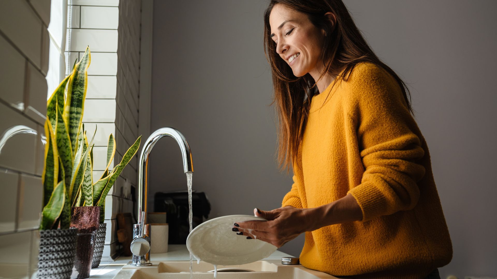 Woman in yellow sweater washes dishes in a bright kitchen, smiling.
