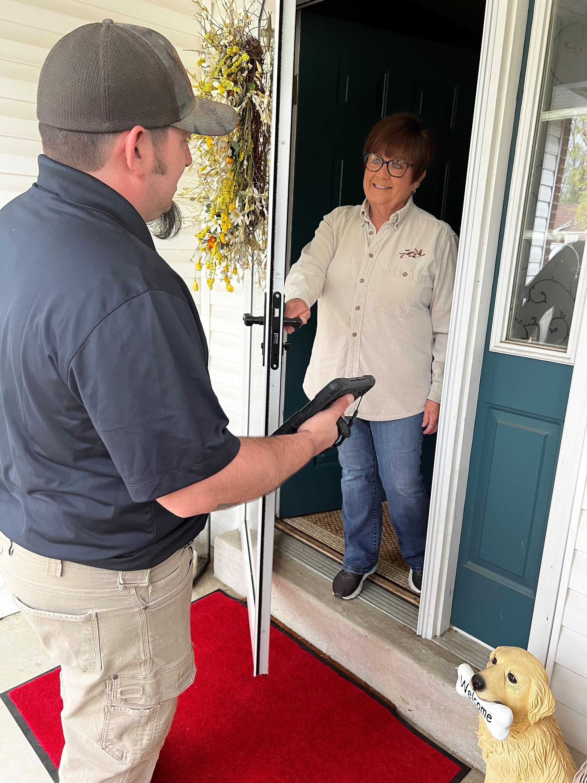 Man handing a device to a woman at a house's door. The woman is smiling. A golden retriever sits on the porch.