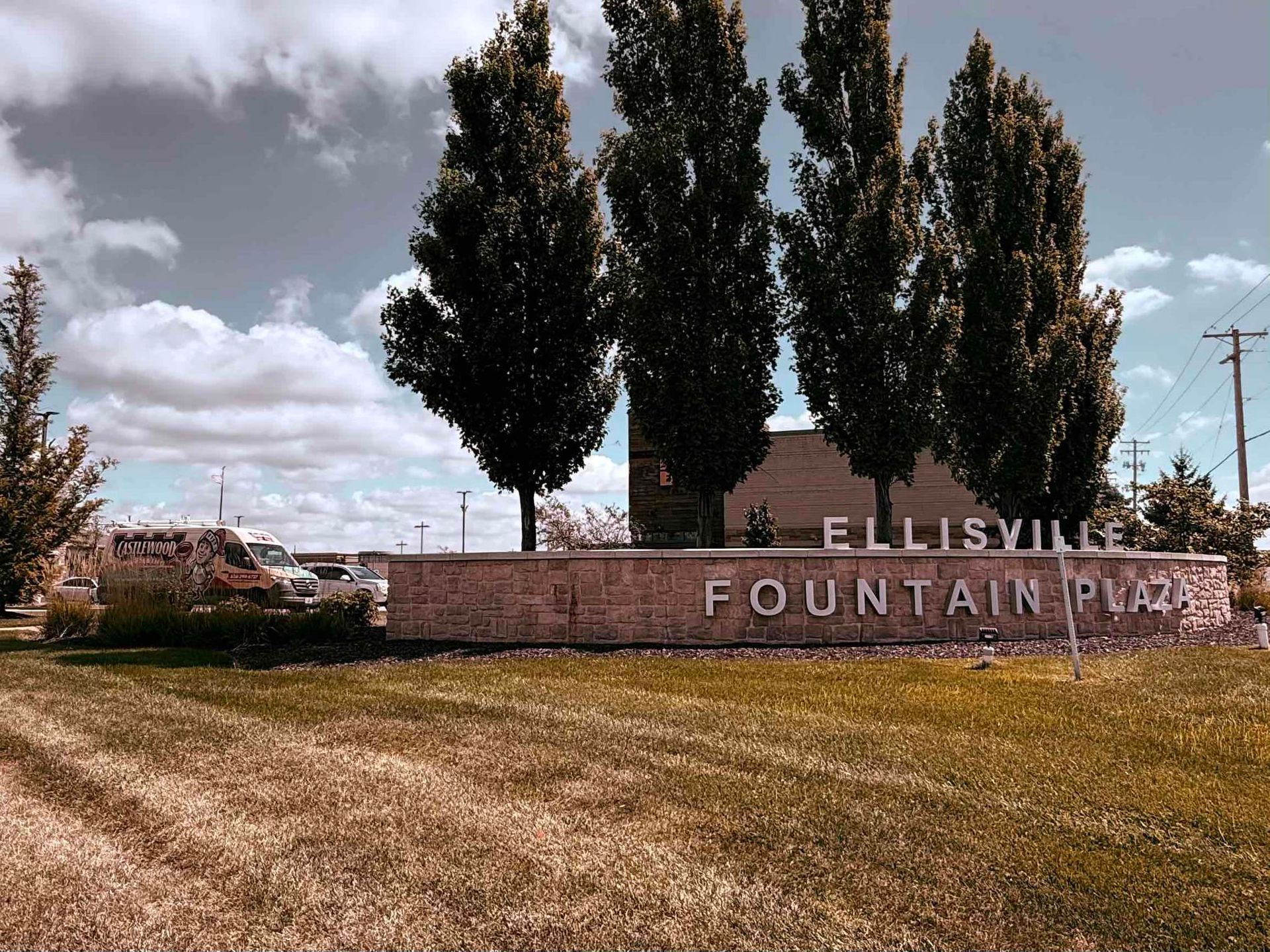 Sign for Ellisville Fountain Plaza with trees and brown grassy field.