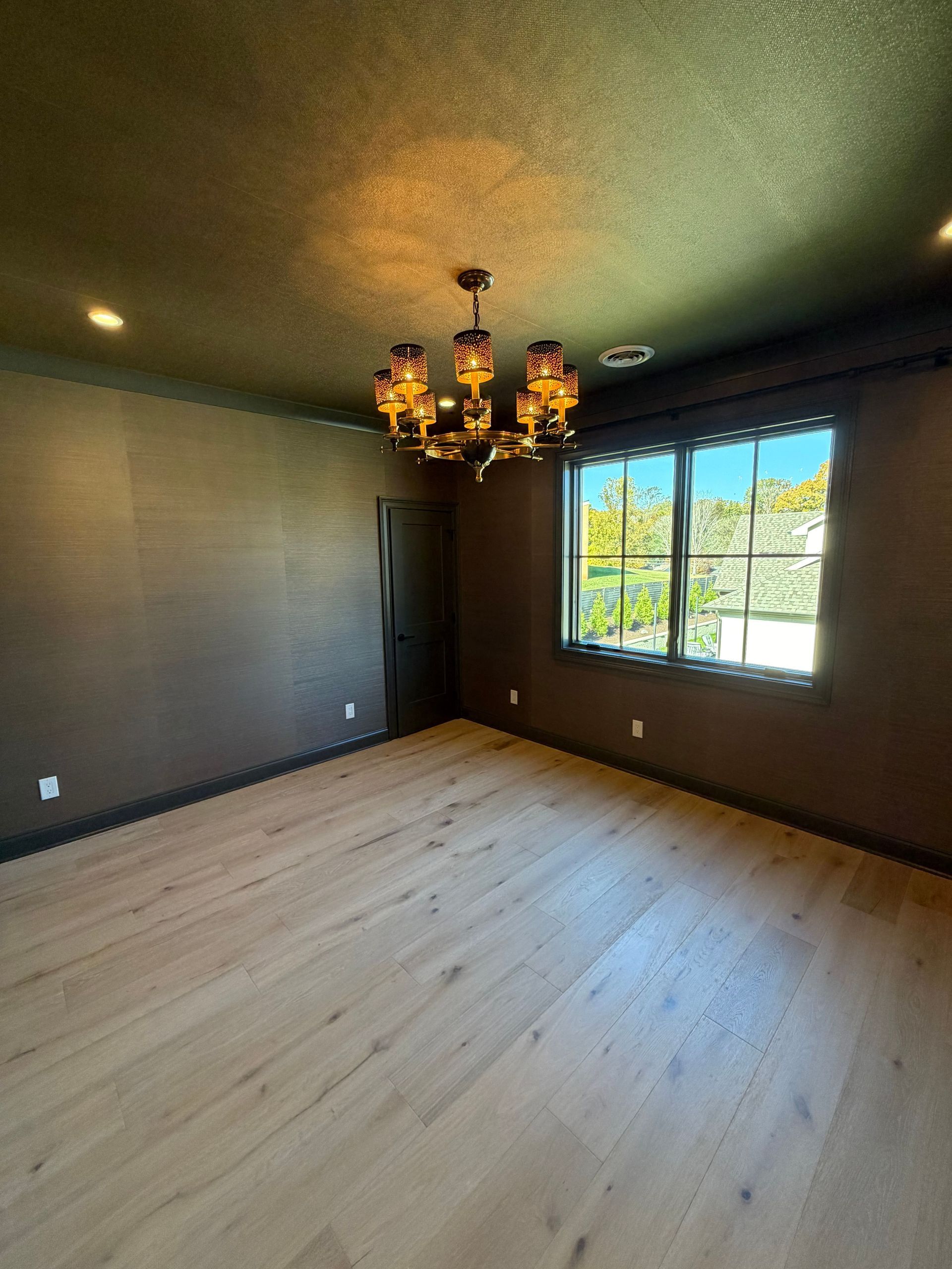 Empty room with dark walls, light wood floor, chandelier, and a window letting in daylight.