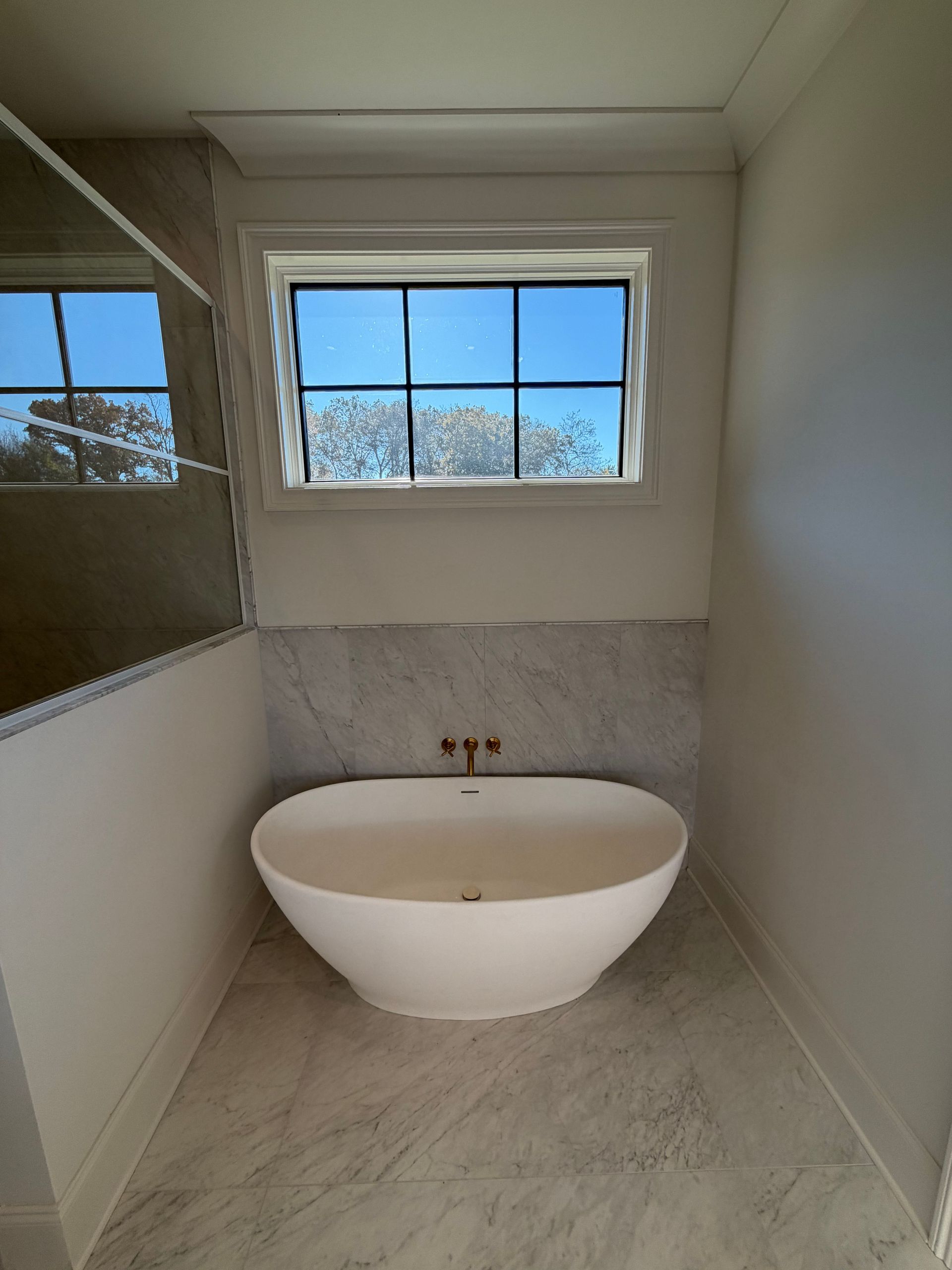 Minimal bathroom with a freestanding tub, large window, and light stone flooring.