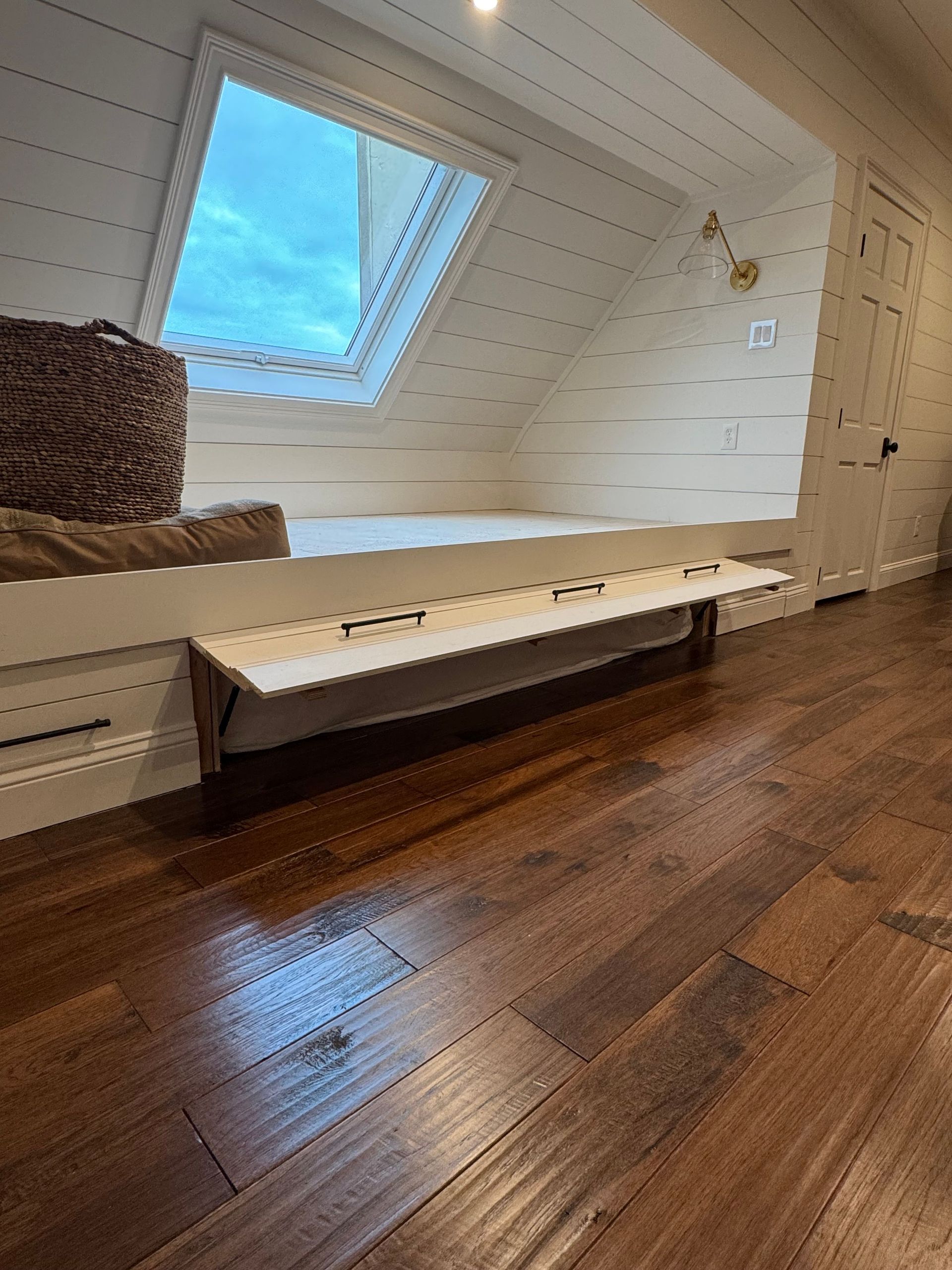 Bright attic room with a skylight, white paneled walls, wooden floor, and a built-in bench.