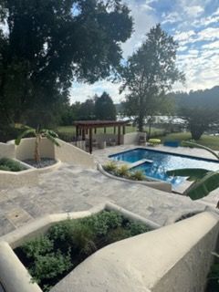 Poolside garden with blue water, white stone paths, and large trees in the background