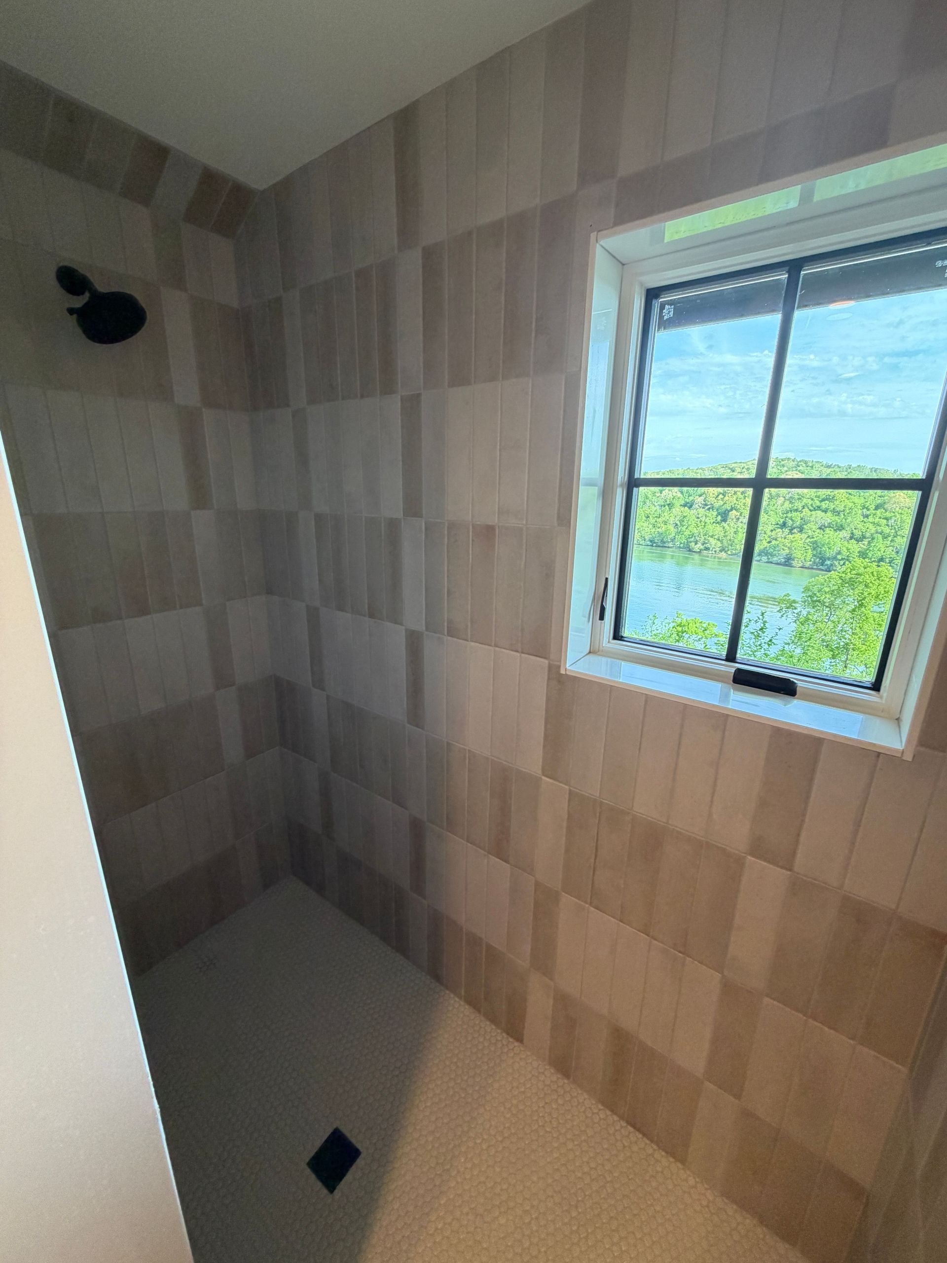 Empty tiled shower with a small window and black floor drain, lit by daylight.