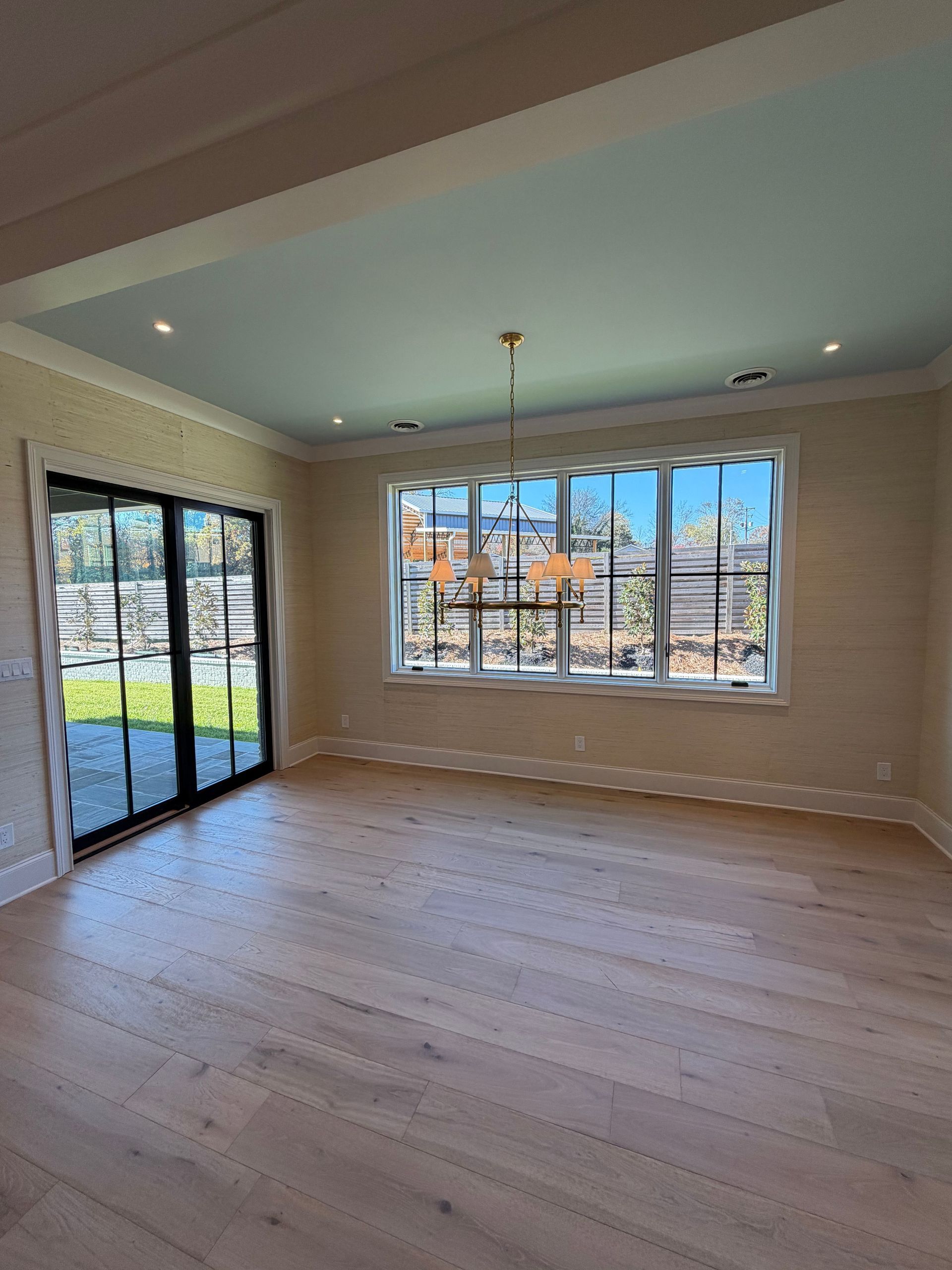Empty sunlit room with hardwood floors, large windows, and sliding glass doors.