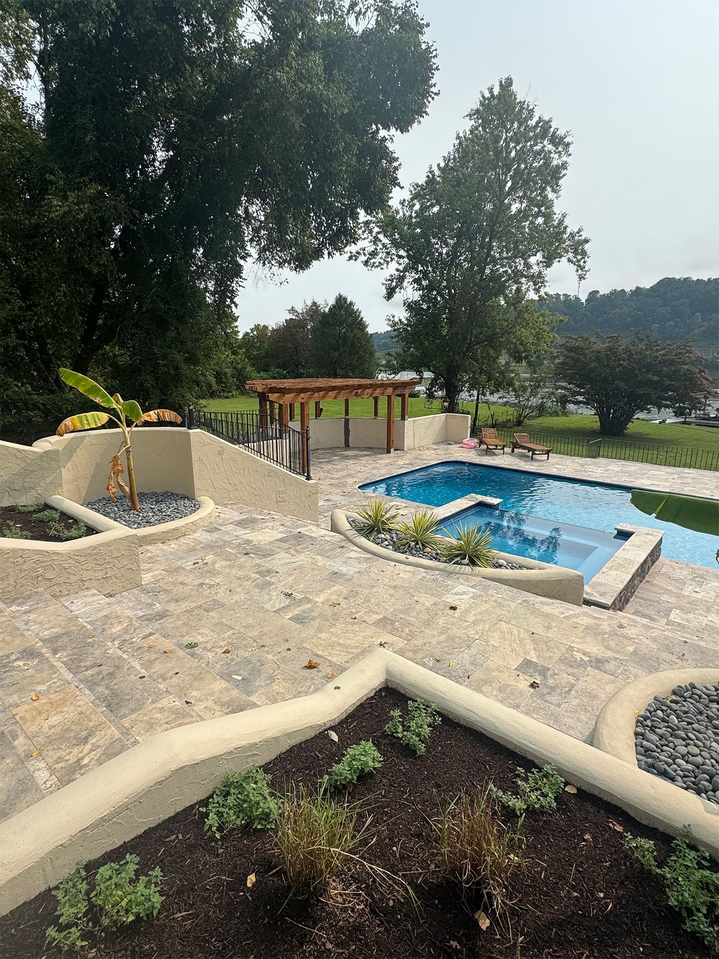 Stone garden terraces beside a blue pool, with trees, a pergola, and distant hills.