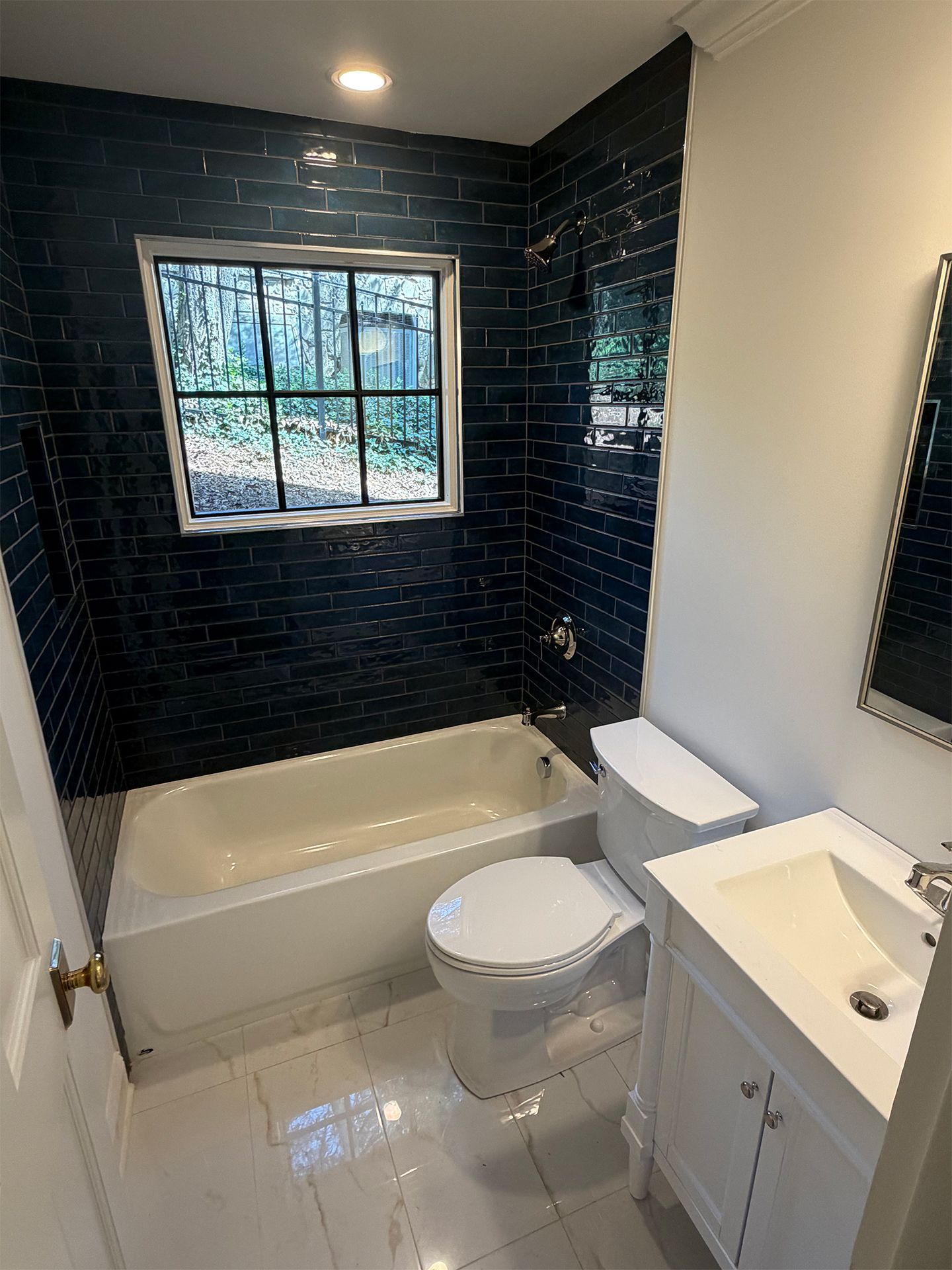 Small bathroom with black tile walls, white tub, toilet, and vanity by a window.