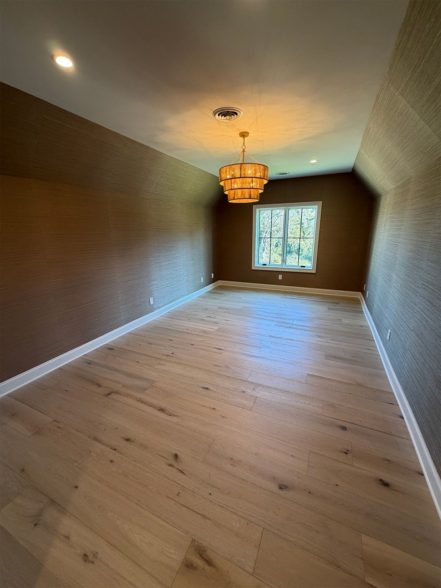 Empty long room with wood floors, brown walls, white trim, ceiling light, and a window at the far end.
