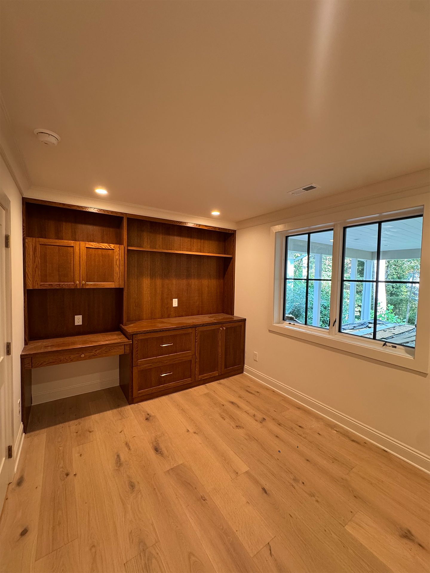Empty wood-paneled room with built-in cabinets, light wood floors, and a window overlooking greenery