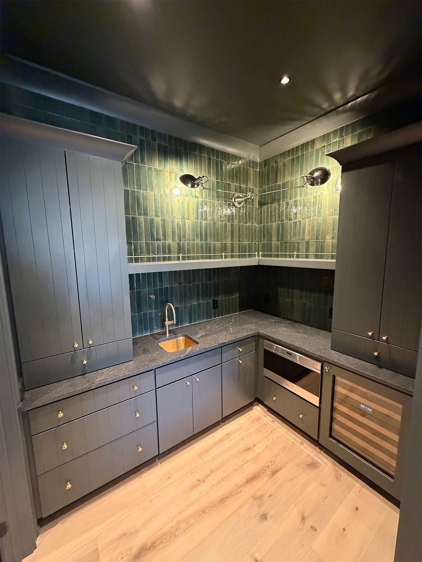 Dark modern kitchen with green tiled backsplash, black cabinets, light wood floor, sink, and built-in oven.