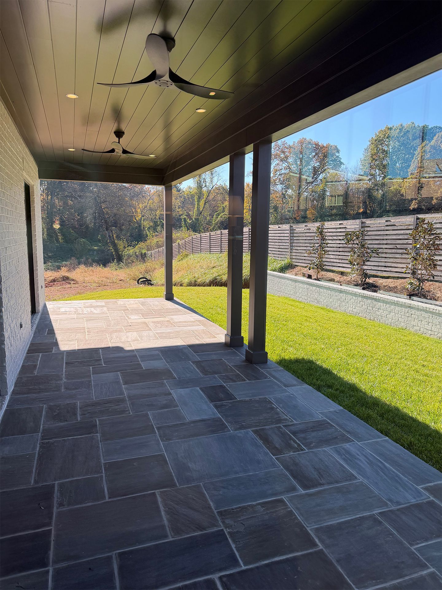 Covered stone patio with ceiling fans, overlooking a sunny lawn and wooden fence