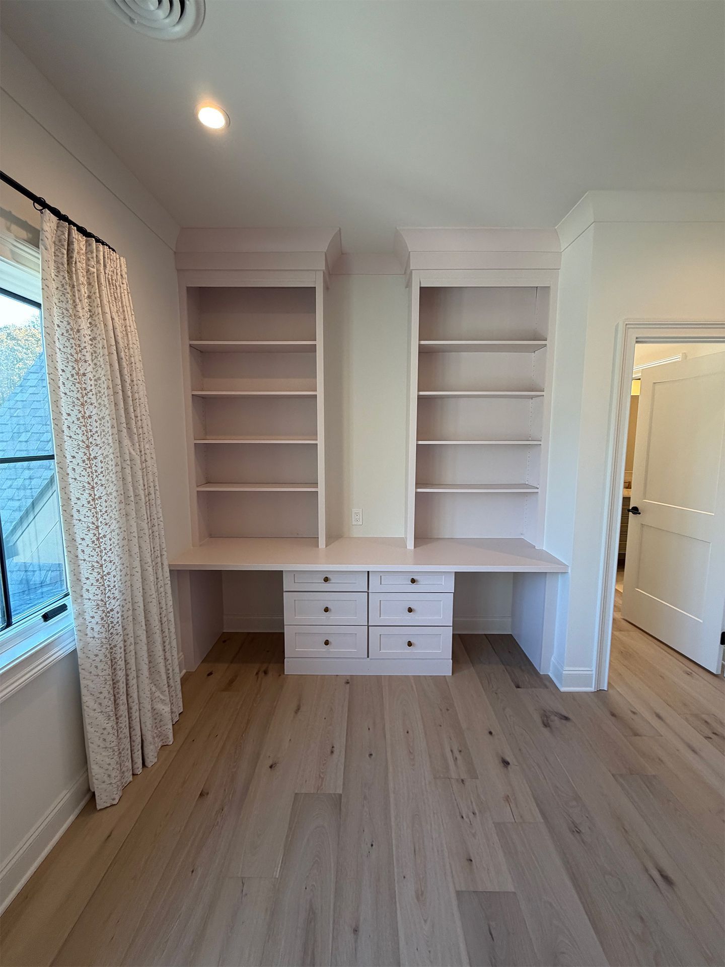 White built-in shelving and desk with drawers in a bright room, light wood floor, window and curtain.