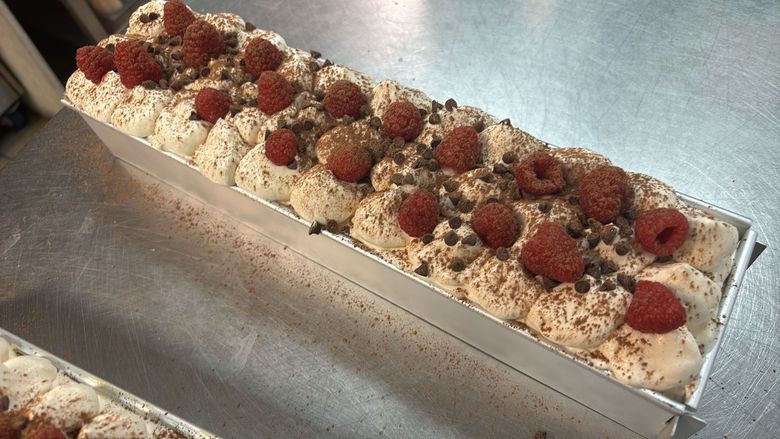 Un gâteau rectangulaire garni de crème fouettée, de framboises fraîches et de copeaux de chocolat, présenté dans un moule en métal.