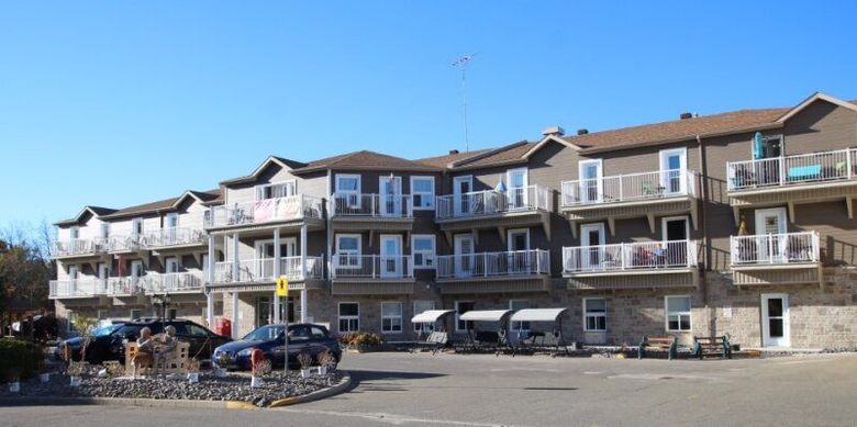Un immeuble à plusieurs étages, avec un revêtement beige, un soubassement en pierre et des rangées de balcons blancs, sous un ciel bleu limpide.