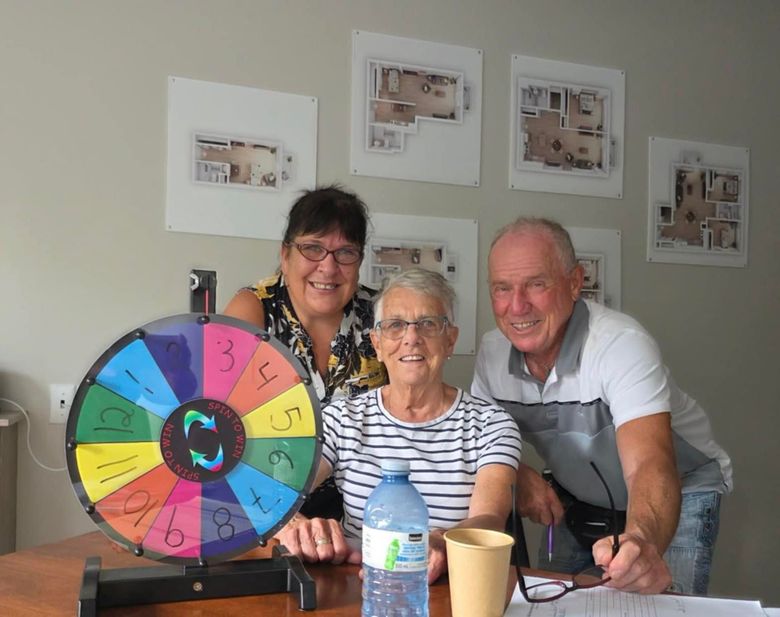 Trois personnes sourient derrière une table où se trouve une roue de la fortune colorée numérotée de 1 à 12, dans une pièce ornée de plans d'étage.