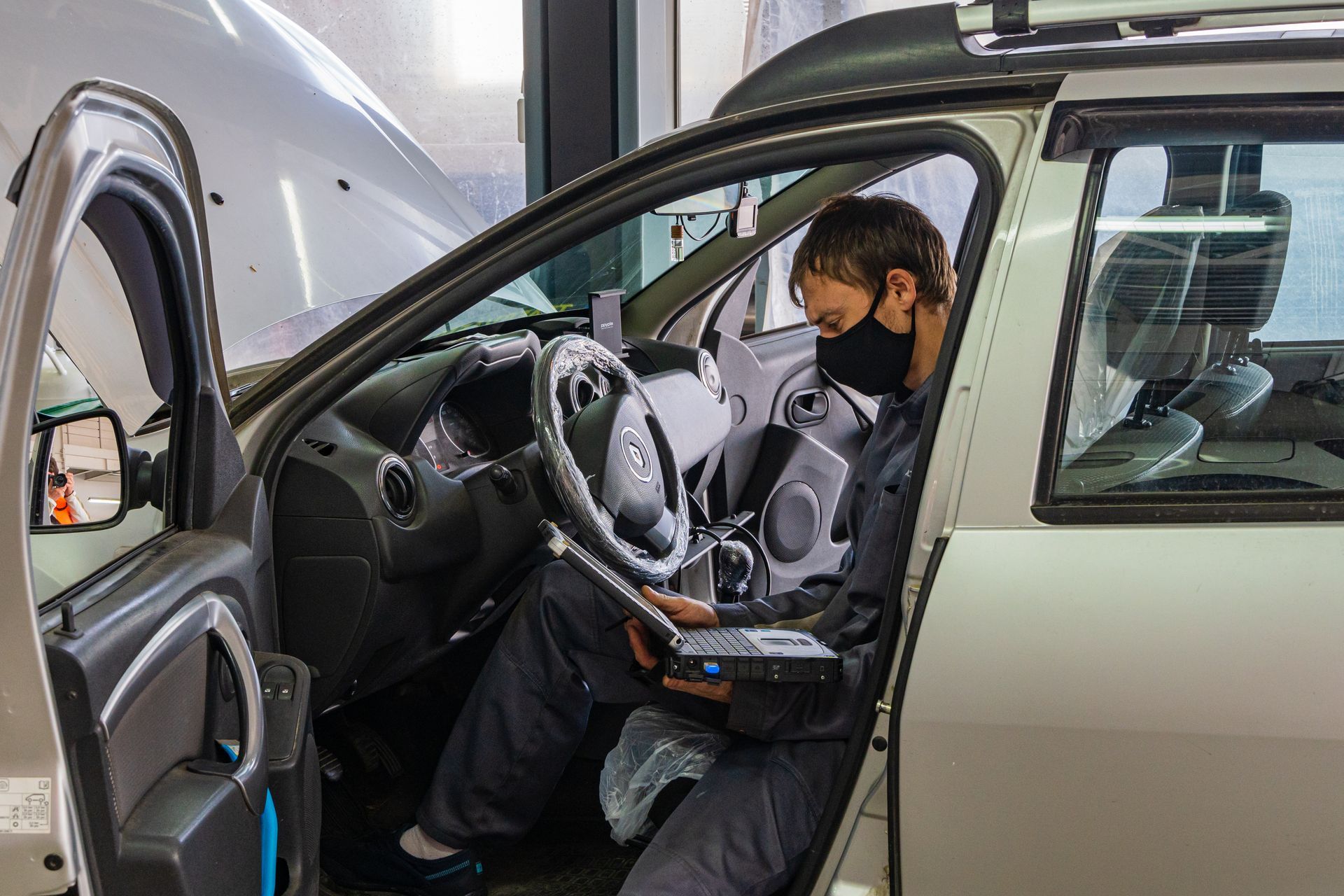 Mechanic in black mask using a diagnostic tool inside a car in a garage.