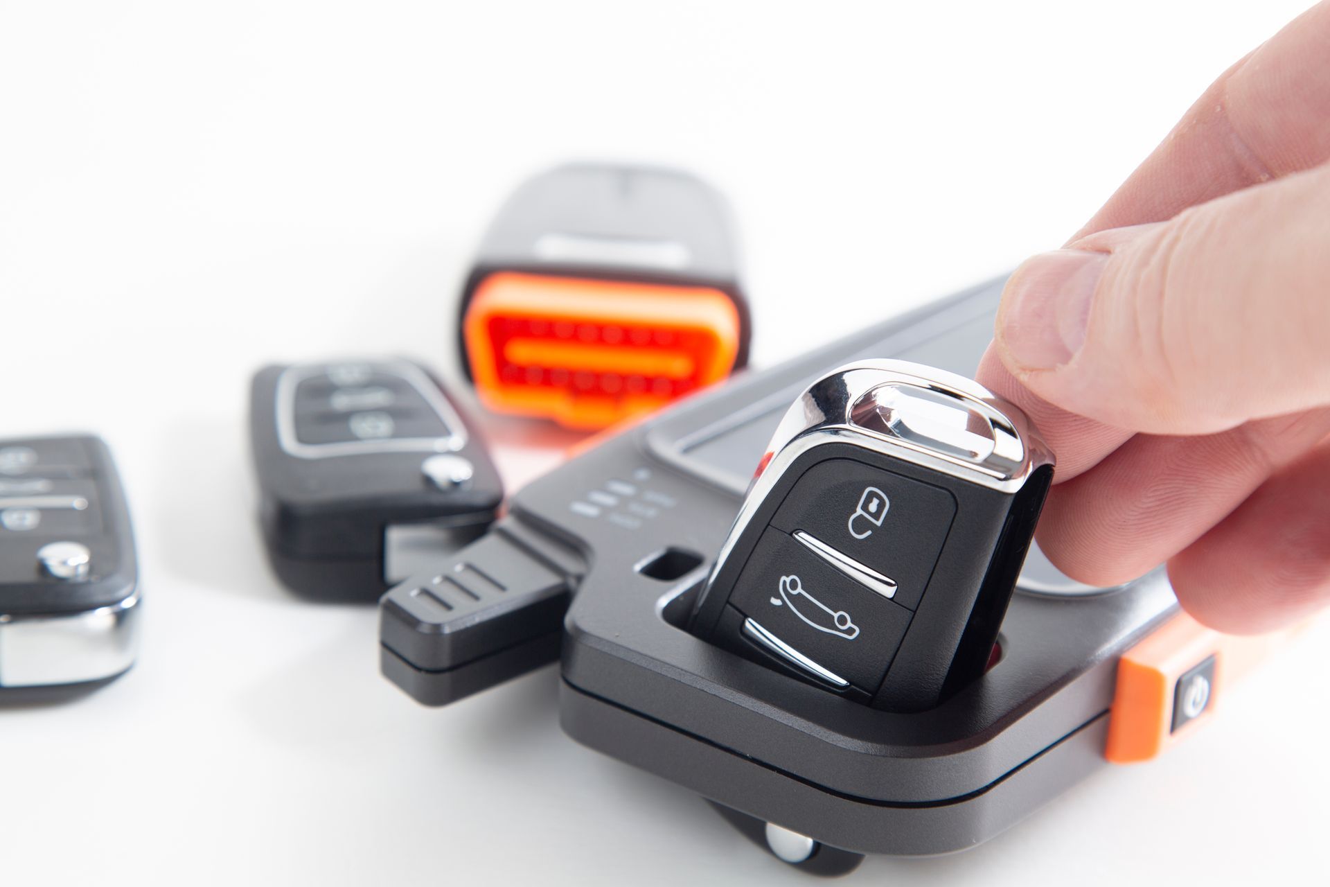 Hand holding car key next to a black programming device; other car keys and a reader are visible.