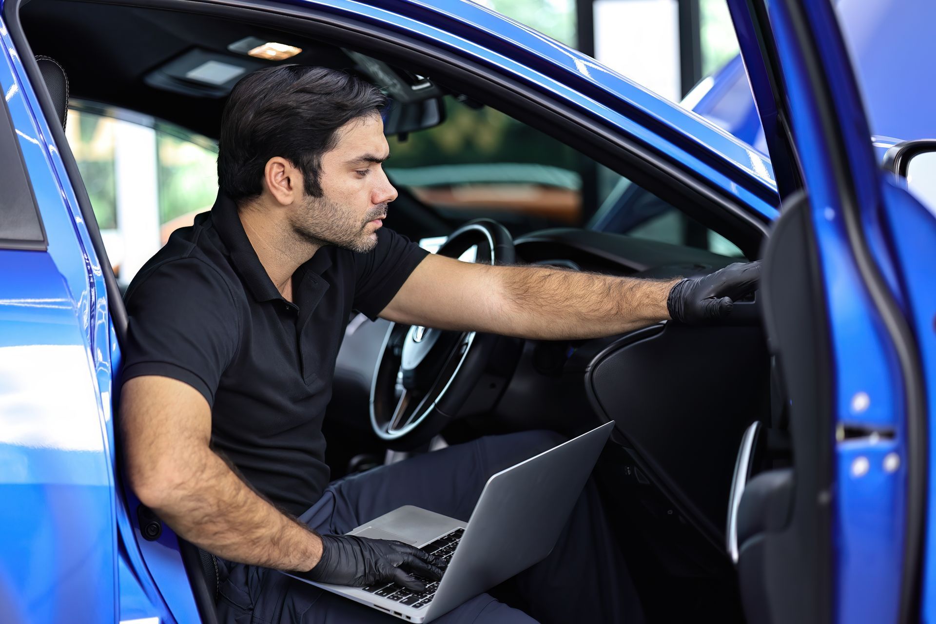 Mechanic in black gloves using laptop to diagnose a blue car's dashboard.