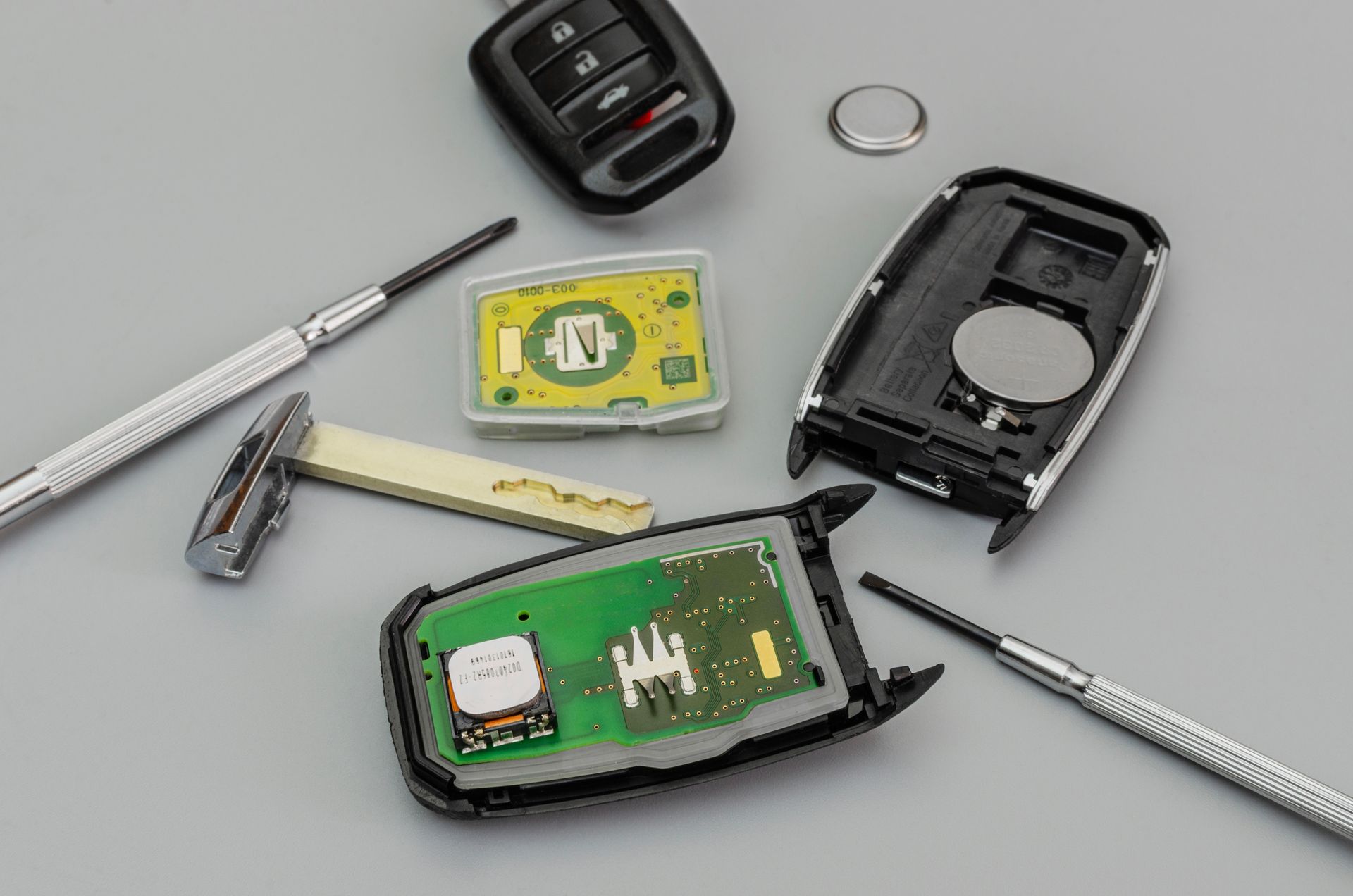 Disassembled car key with circuit board, battery, and tools on a gray surface.