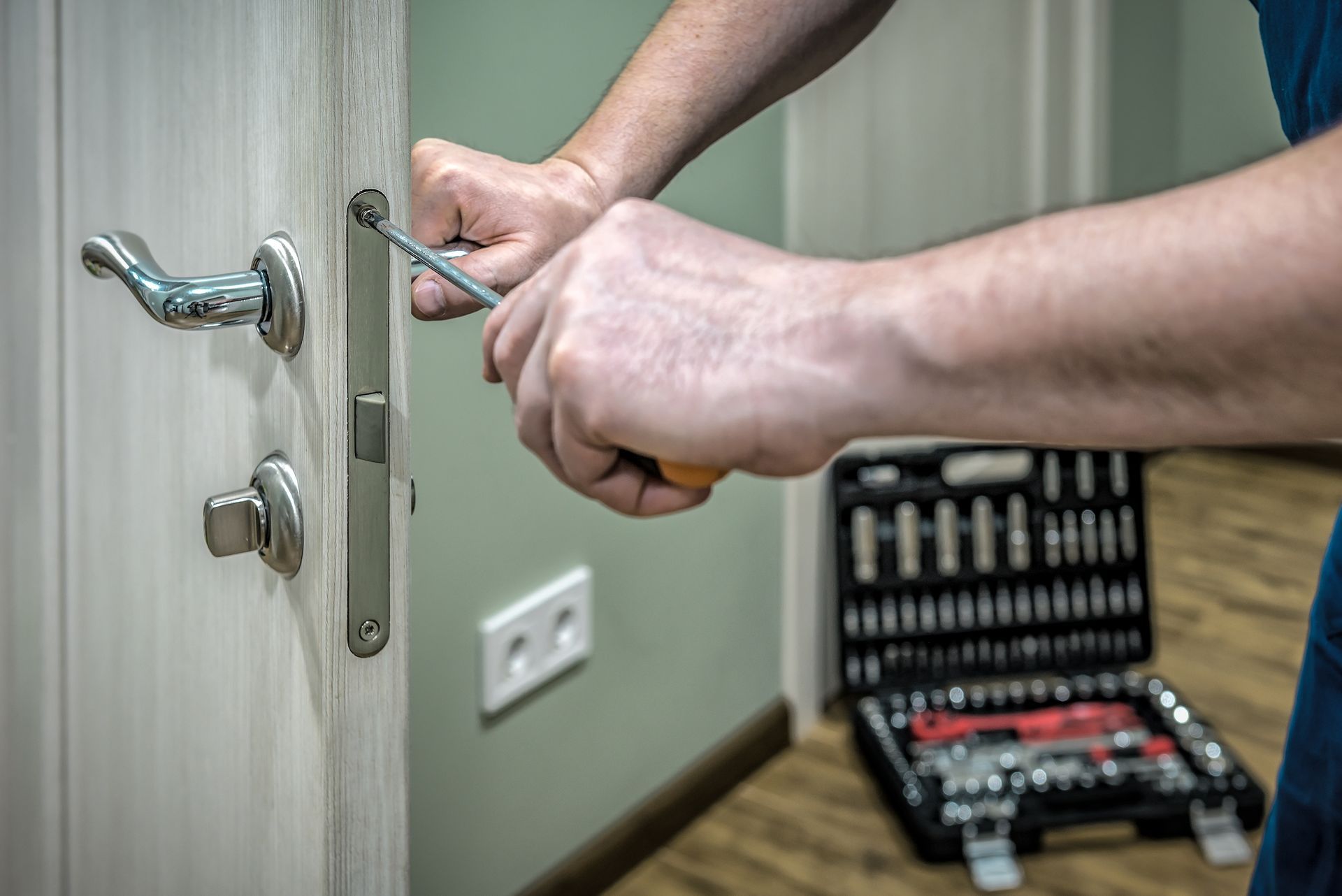 Person uses a screwdriver to fix a door latch on a white door. A toolbox sits on the floor nearby.