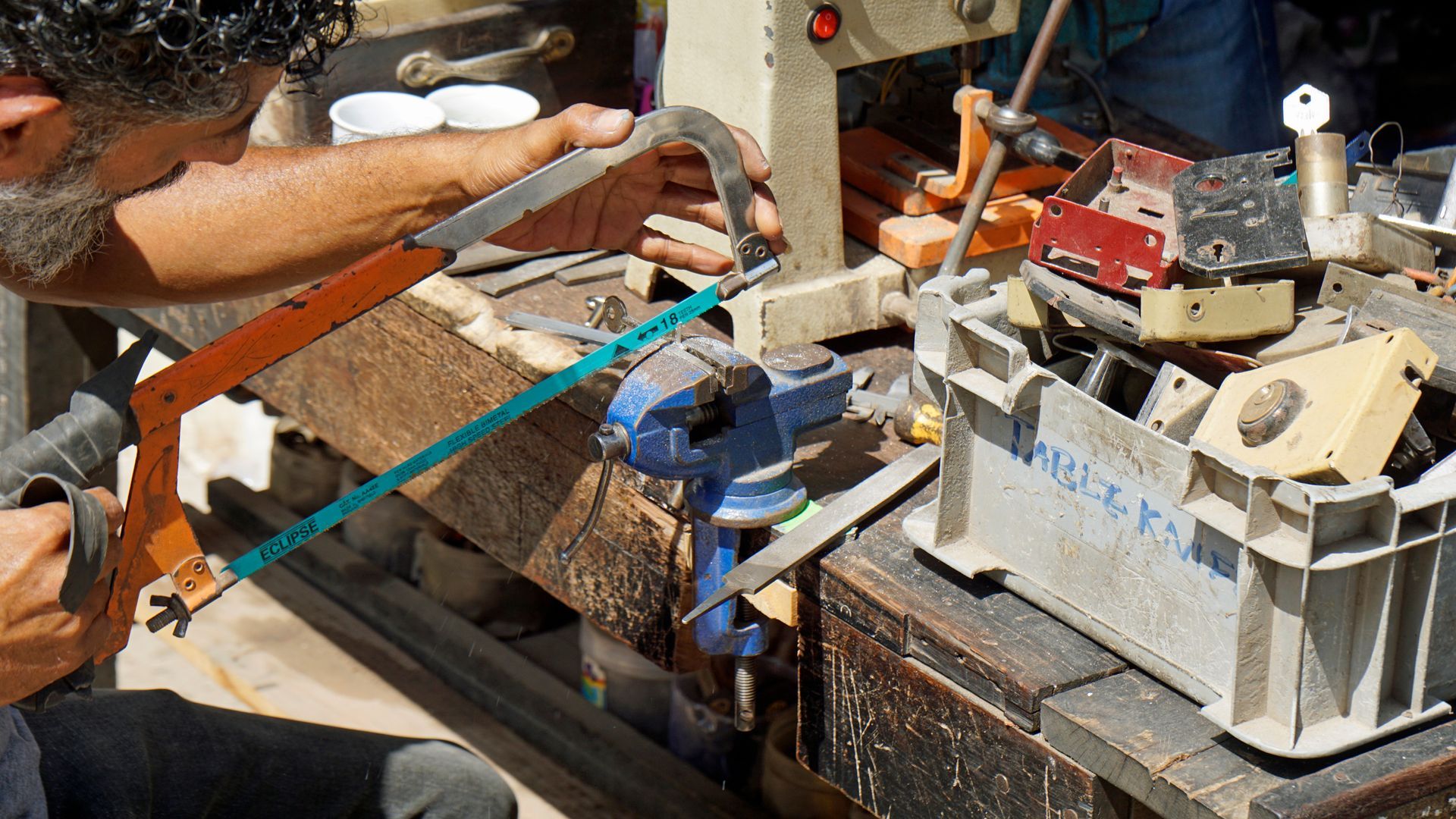 Person sawing wood clamped in a blue vise on a workbench, surrounded by tools and equipment.