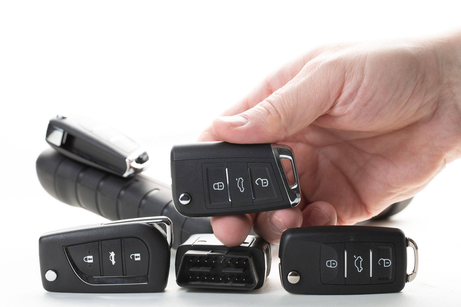 Hand holding a car key fob among several other car keys on a white surface.