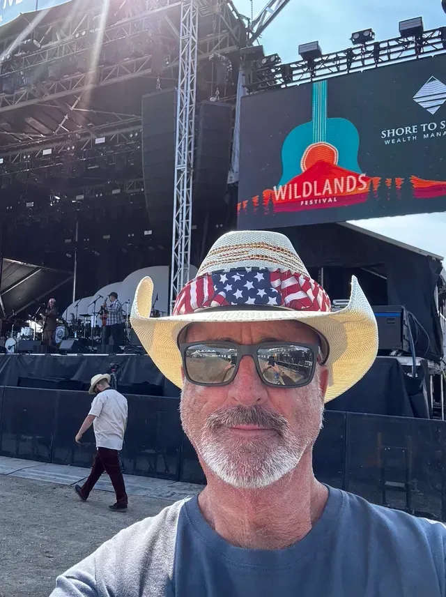 A man wearing a cowboy hat and sunglasses is taking a selfie in front of a stage.
