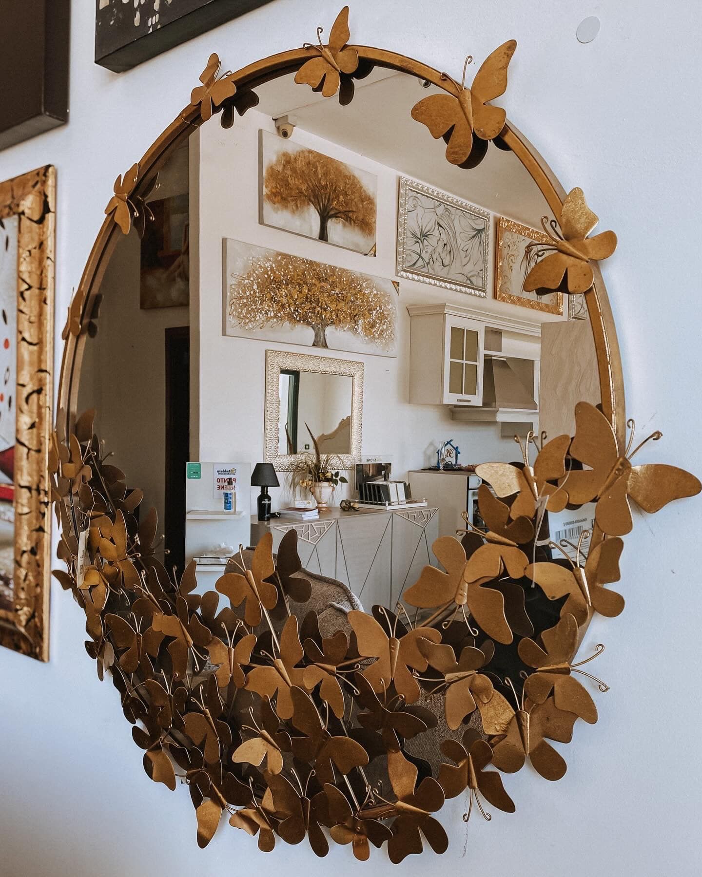 Specchio ovale con decorazioni dorate a forma di farfalla e foglia, che riflette una stanza con opere d'arte e mobili.
