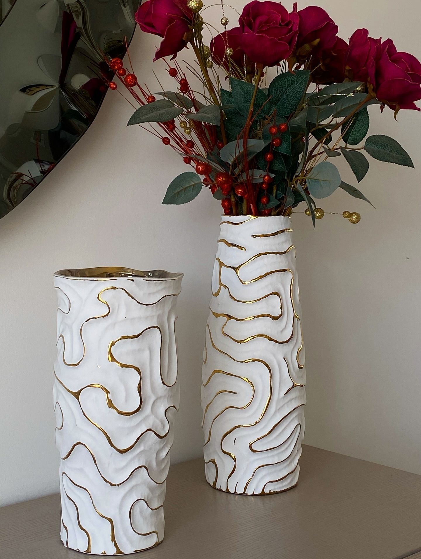 Due vasi bianchi con dettagli dorati, uno dei quali contiene rose rosse e bacche, su una mensola.