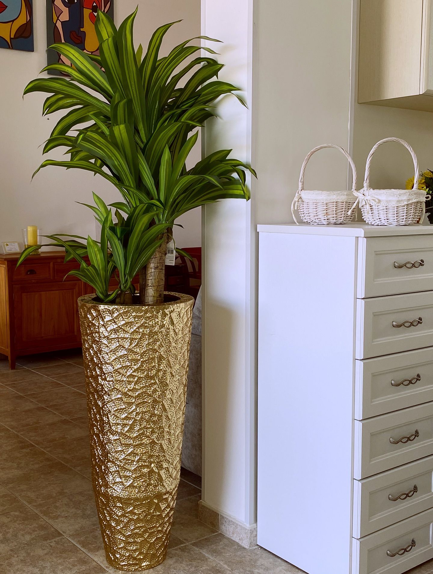 Fioriera alta con texture dorata, con una grande pianta verde, accanto a un mobiletto bianco e due piccoli cestini.