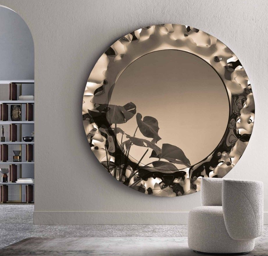 Un grande specchio rotondo e strutturato è appeso a una parete, con delle piante che si riflettono. Di fronte c'è una sedia bianca.