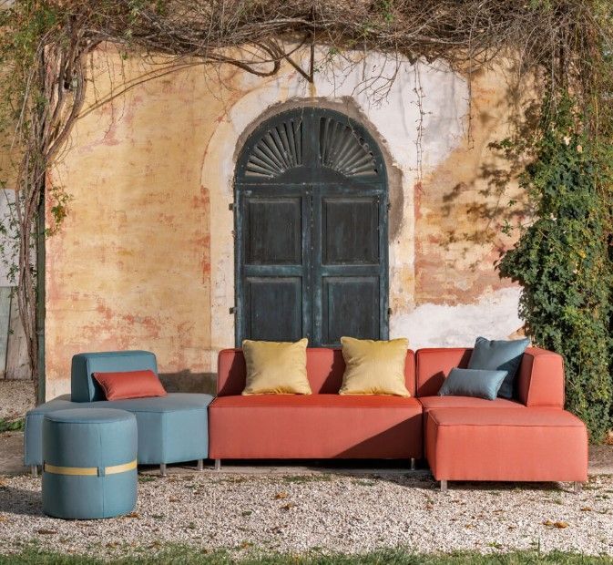 Divano componibile da esterno con tappezzeria turchese e corallo, cuscini gialli e blu, contro un muro e una porta rovinati dal tempo.