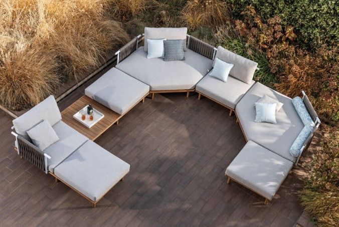 Divano componibile da esterno con cuscini beige, guanciali e tavolino su una terrazza in legno, circondato da piante secche.