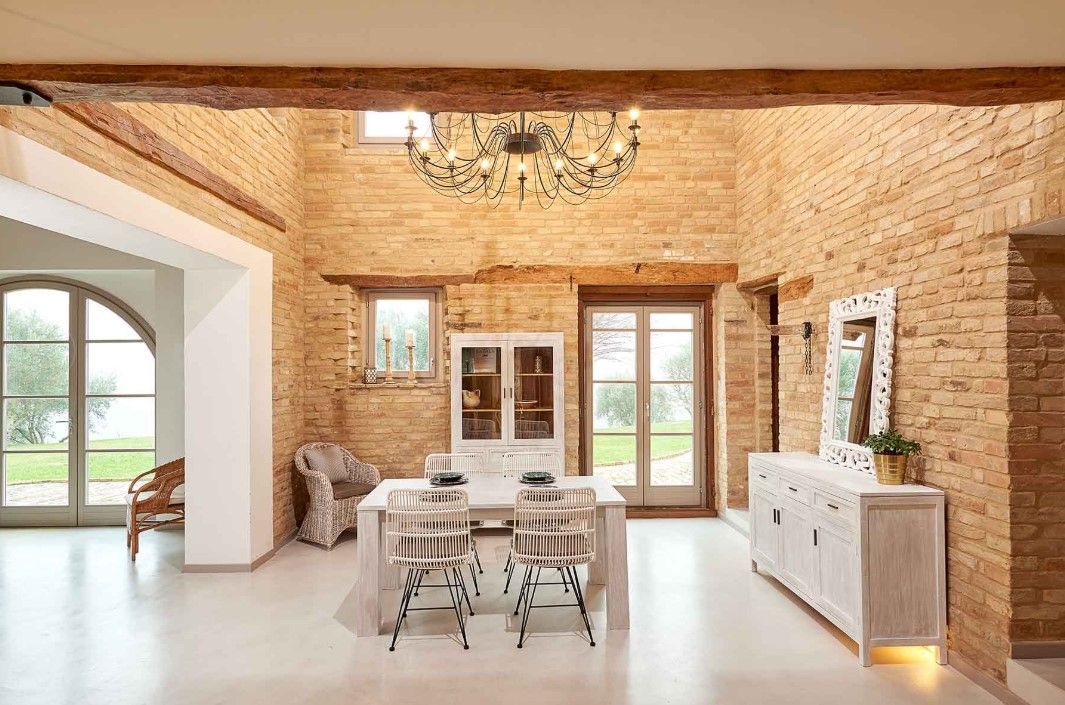 Sala da pranzo con pareti in mattoni a vista, mobili bianchi, lampadario e finestra ad arco con vista.