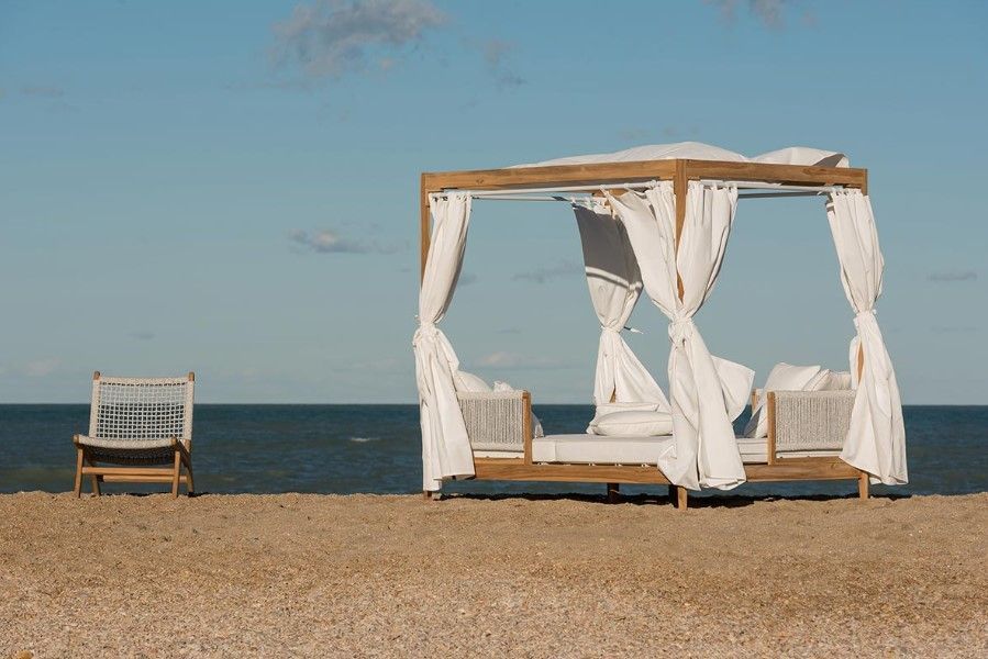 Una cabina da spiaggia con tende bianche è situata su una riva sabbiosa accanto a una sedia, con l'oceano sullo sfondo.