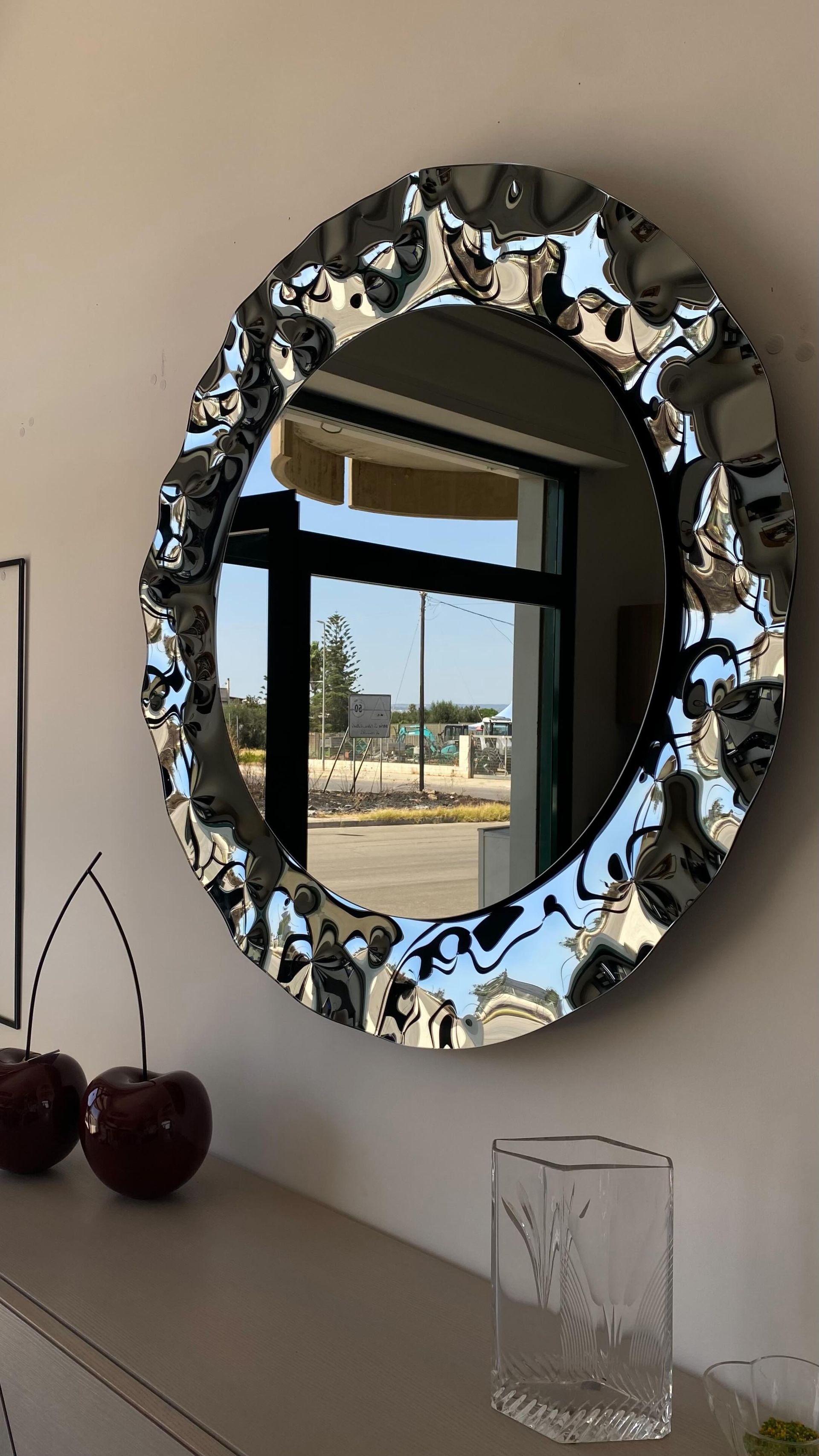 Specchio rotondo con cornice cromata ondulata che riflette la vista esterna. Ciliegie e vaso di vetro sotto.