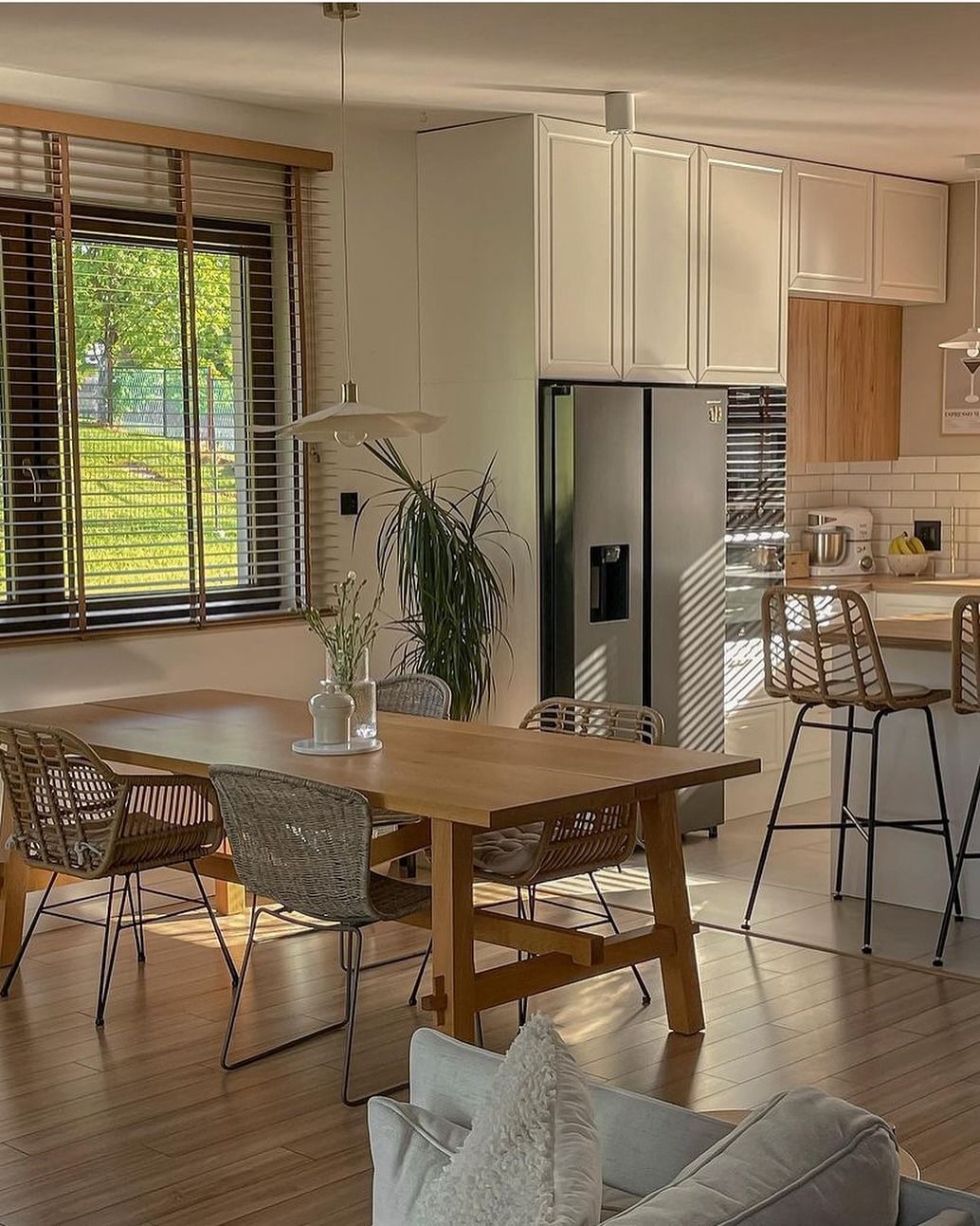 Zona pranzo con tavolo in legno e sedie intrecciate, cucina con mobili bianchi e frigorifero in acciaio inox.