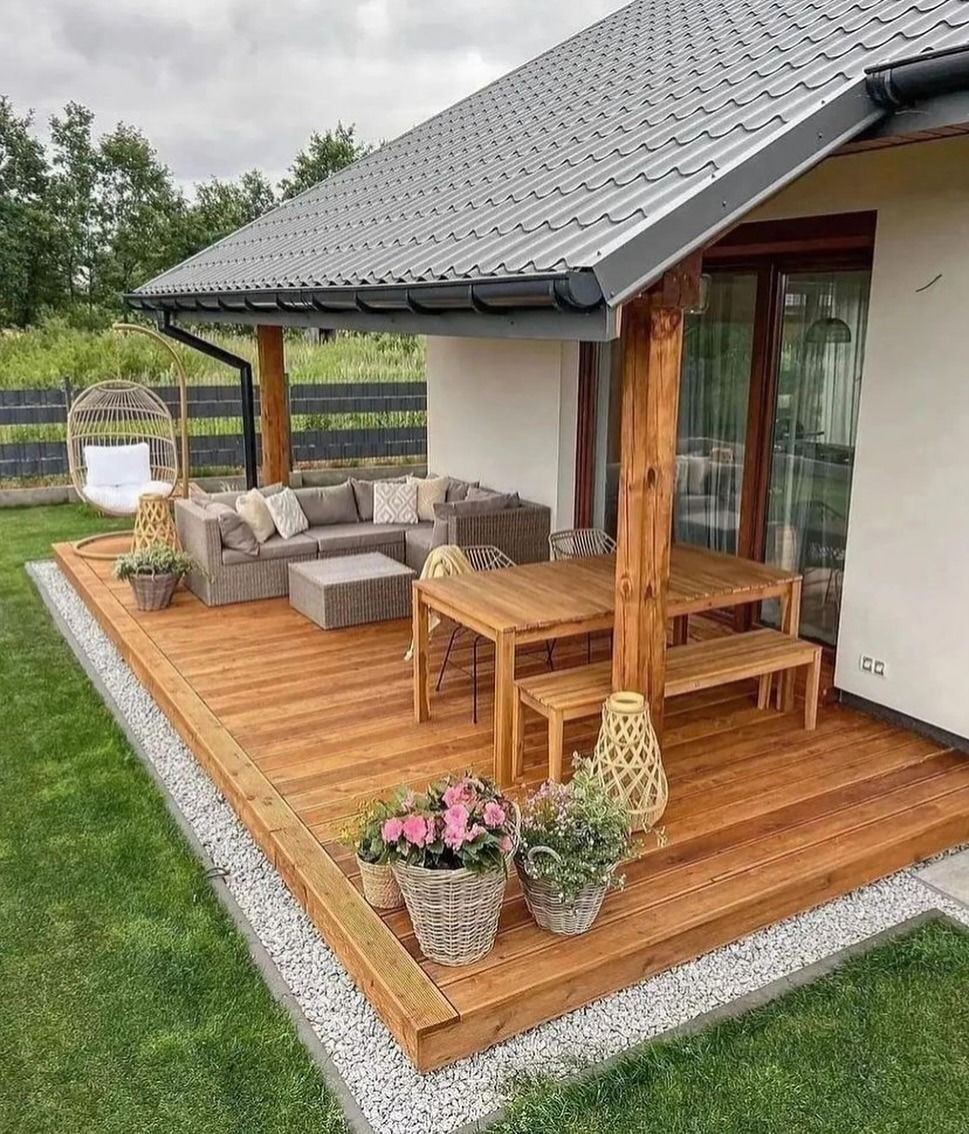 Terrazza in legno con mobili da esterno, tra cui divano, tavolo e panca, accanto a una casa con il tetto grigio.