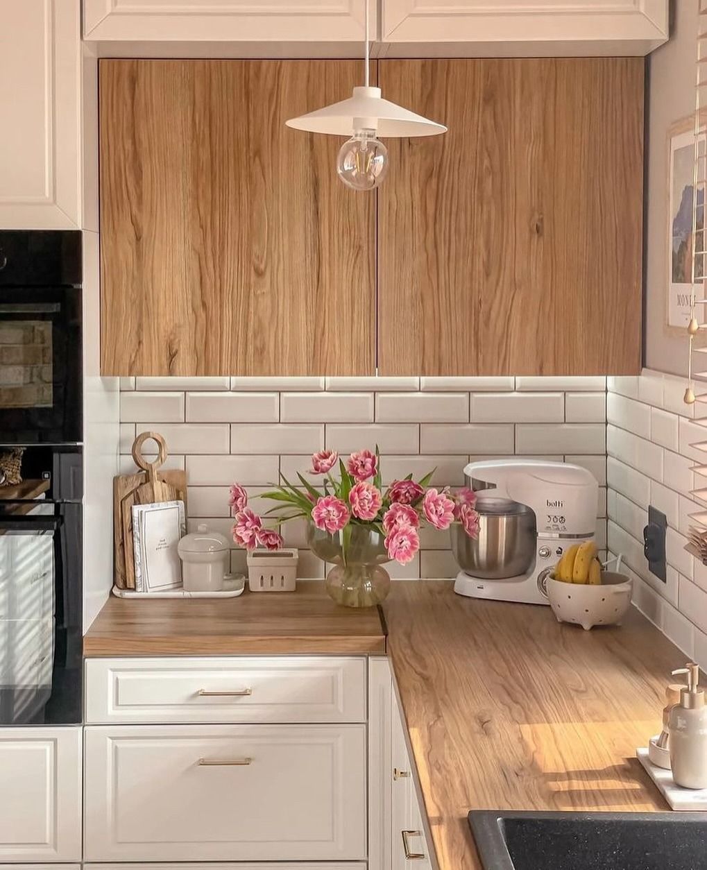 Cucina con mobili in legno, ripiani, paraschizzi in piastrelle bianche della metropolitana, tulipani rosa e lampada a sospensione.