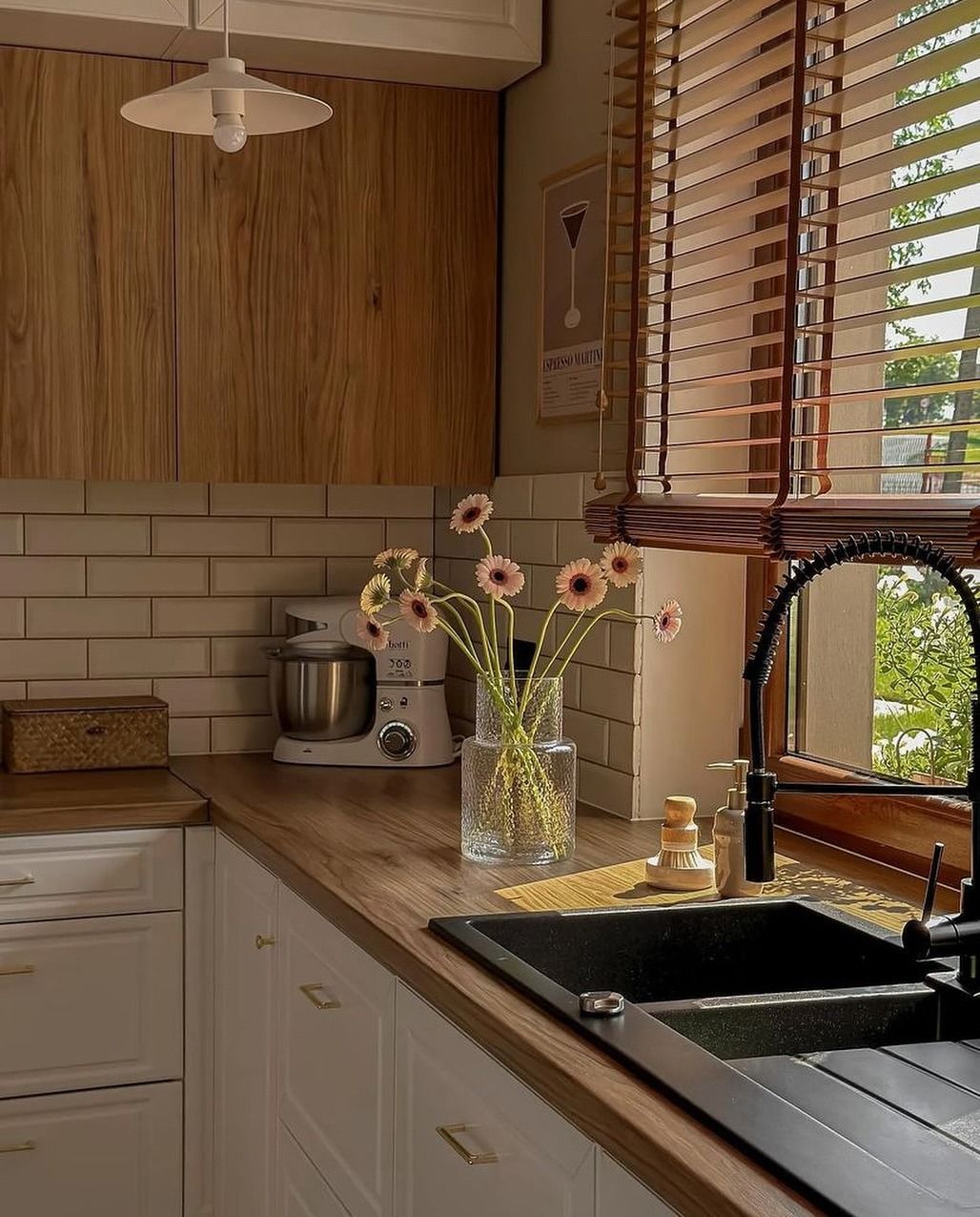 Accogliente cucina con mobili in legno, piano di lavoro e persiane. Fiori in un vaso, lavello nero e miscelatore.
