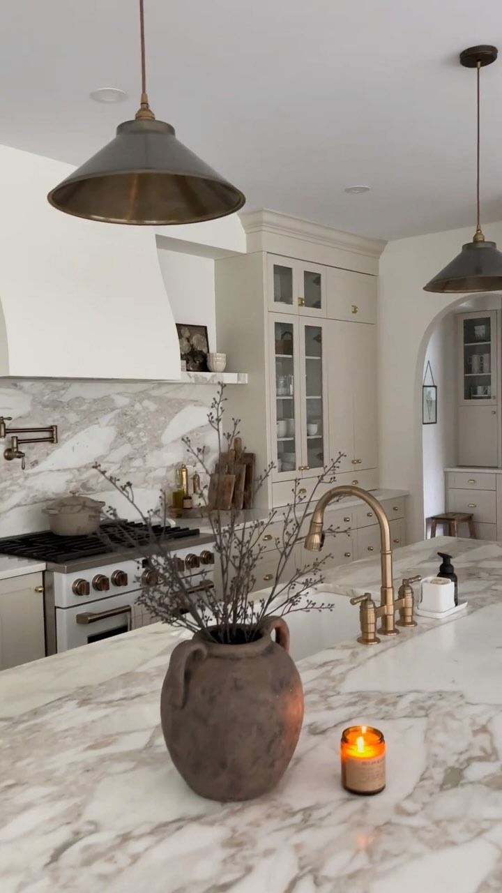 Elegante cucina con ripiani in marmo, infissi in ottone e lampade a sospensione. Un vaso di rami e una candela sono appoggiati sull'isola.
