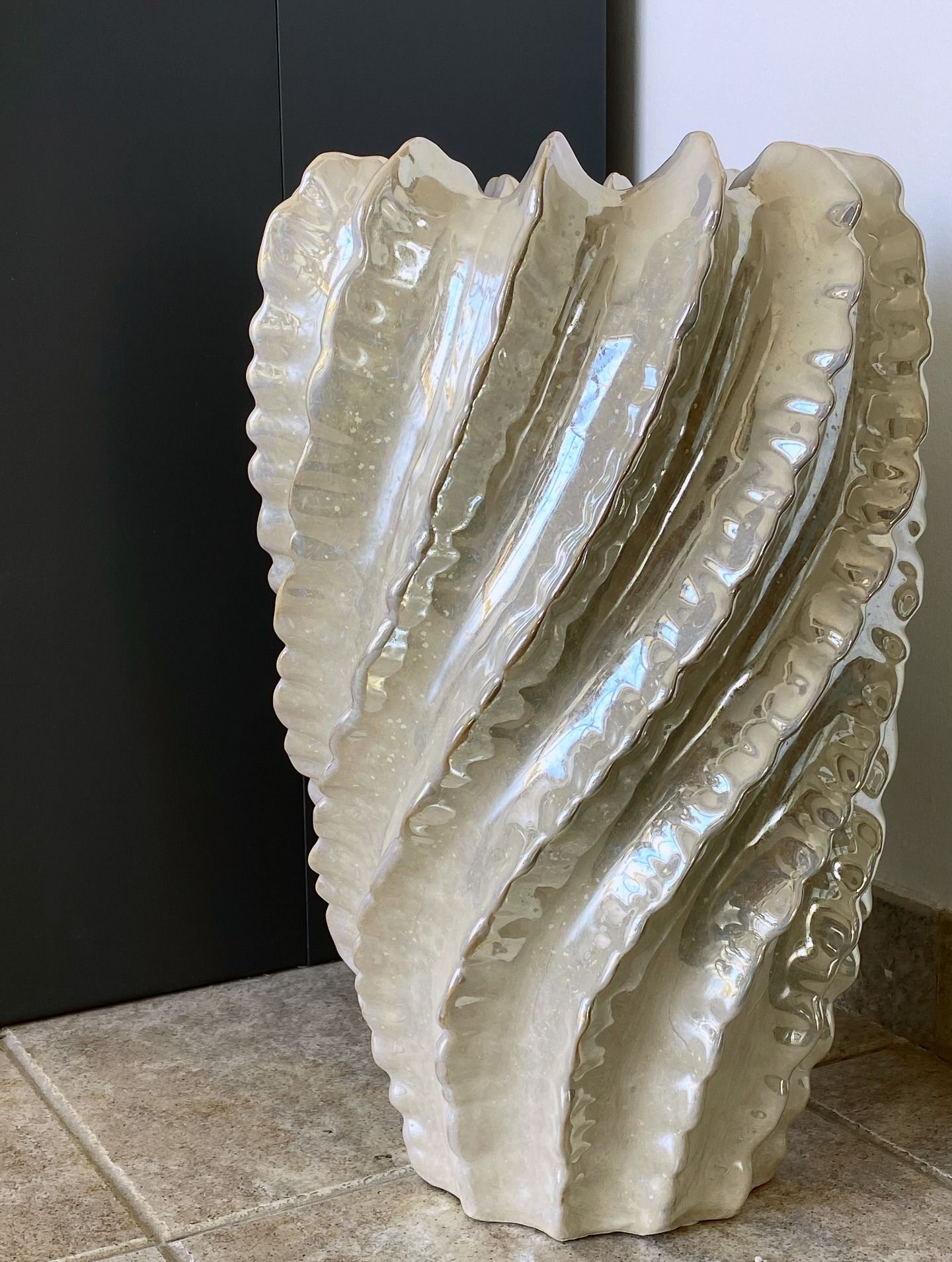 Grande vaso in ceramica bianco sporco, strutturato e ondulato. Appoggiato a una parete grigio scuro e a un pavimento piastrellato.