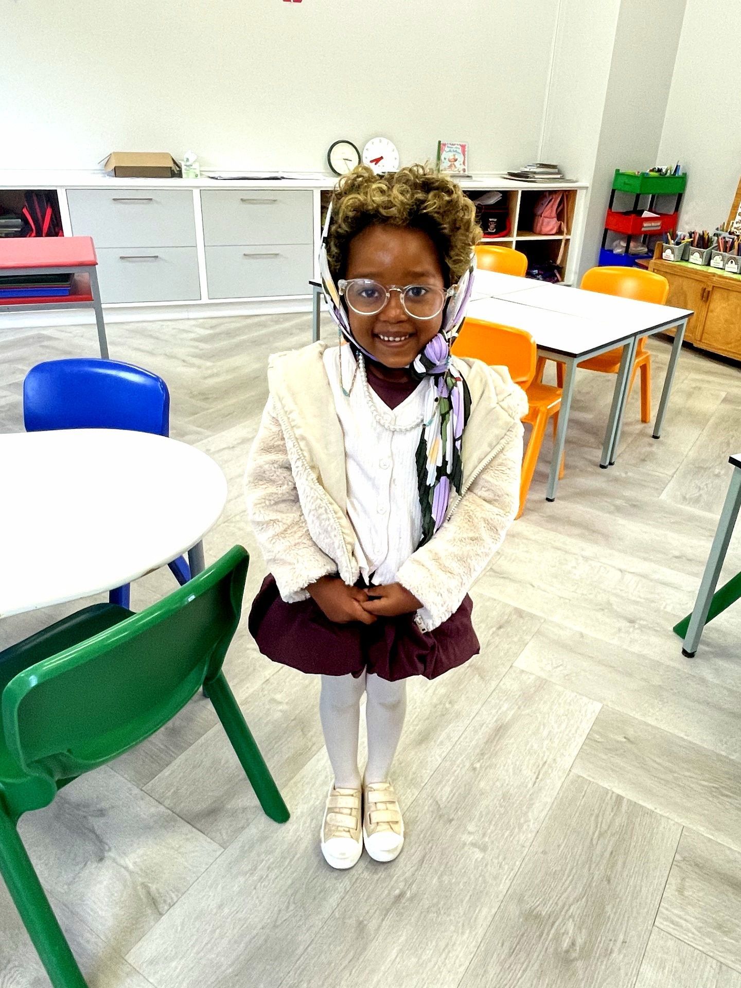 Girl in costume: glasses, scarf, cream jacket, maroon skirt, white tights, gold shoes, smiles in a classroom.