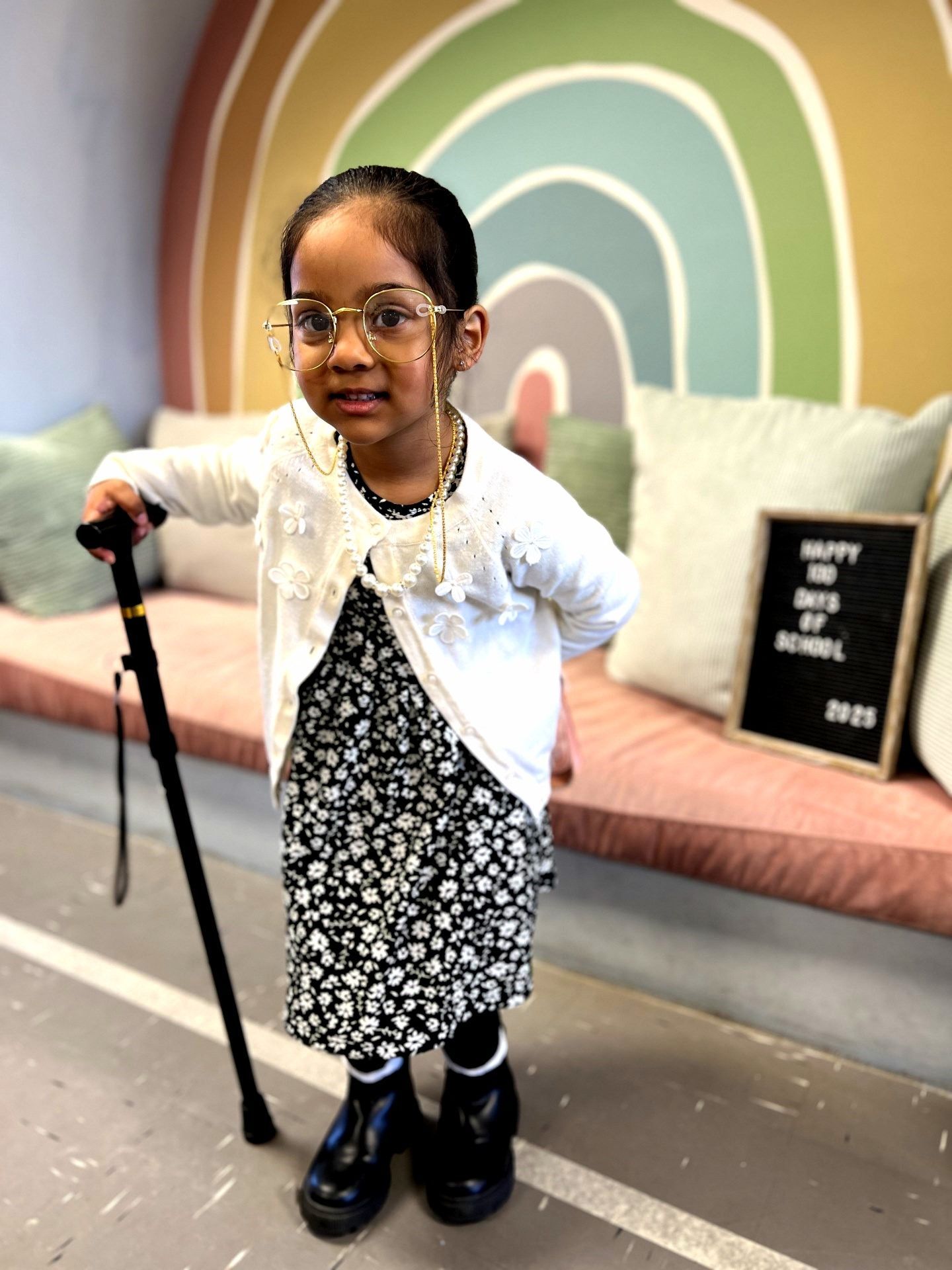 Child dressed as an elderly person for 100th day of school, holding a cane, wearing glasses, in front of rainbow decor.